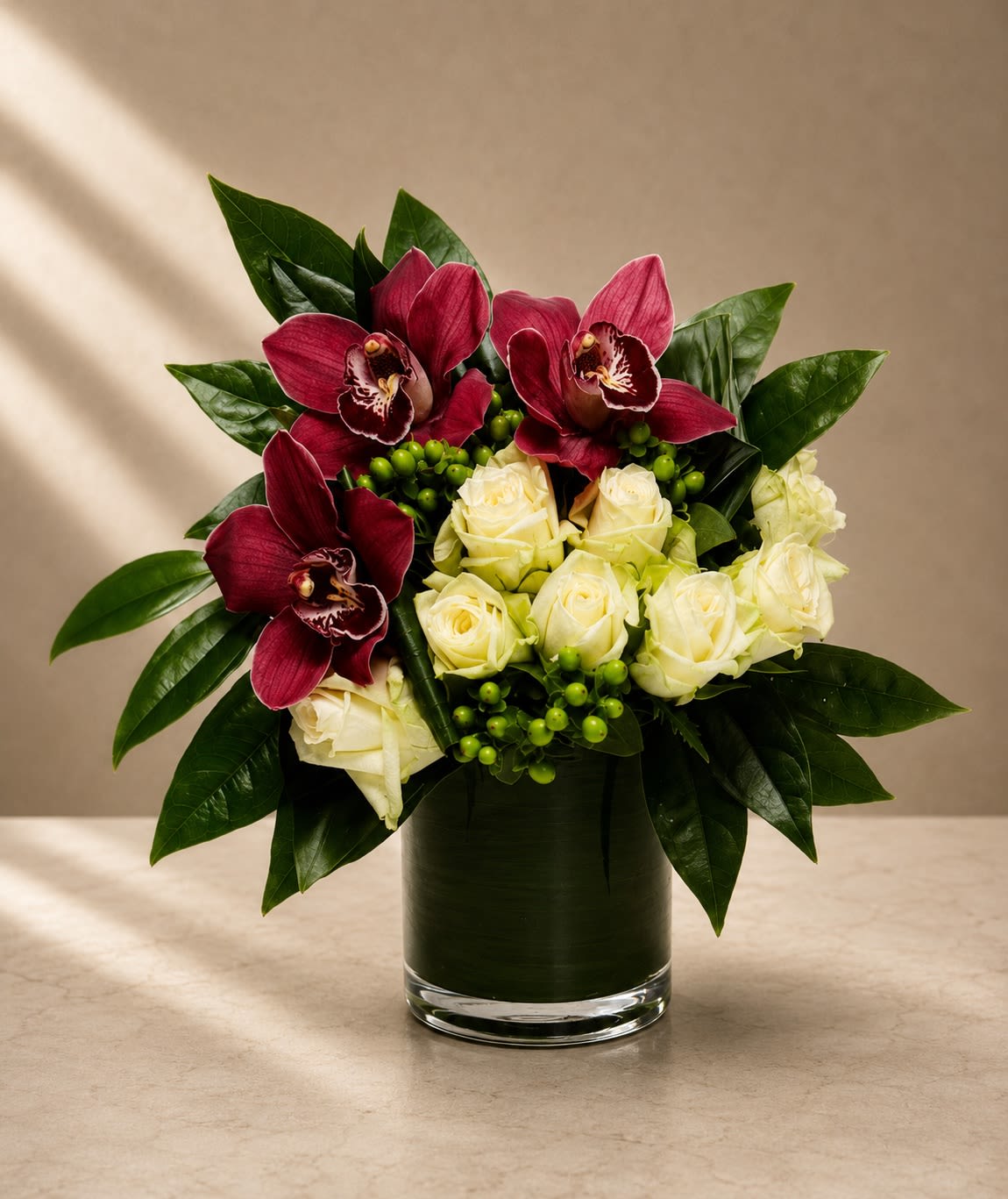 Noir Orchid Elegance - Sophisticated, dramatic, and undeniably luxurious, Noir Orchid Elegance is a couture-inspired floral design that blends deep jewel tones with timeless white roses for a striking modern statement.  This arrangement features rich burgundy cymbidium orchids paired with premium ivory roses and vibrant green hypericum berries, all framed with glossy tropical foliage in a sleek leaf-lined glass cylinder vase. The contrast between the velvety dark orchids and soft creamy roses creates a refined, upscale aesthetic with a bold contemporary edge.  The orchids bring an exotic, fashion-forward presence while the white roses soften the design with classic romance and elegance. Designed with balance and clean structure, this arrangement feels polished, luxurious, and perfect for high-end gifting.  Ideal for anniversaries, luxury celebrations, corporate gifting, romantic occasions, modern home décor, or upscale sympathy arrangements with an artistic touch.