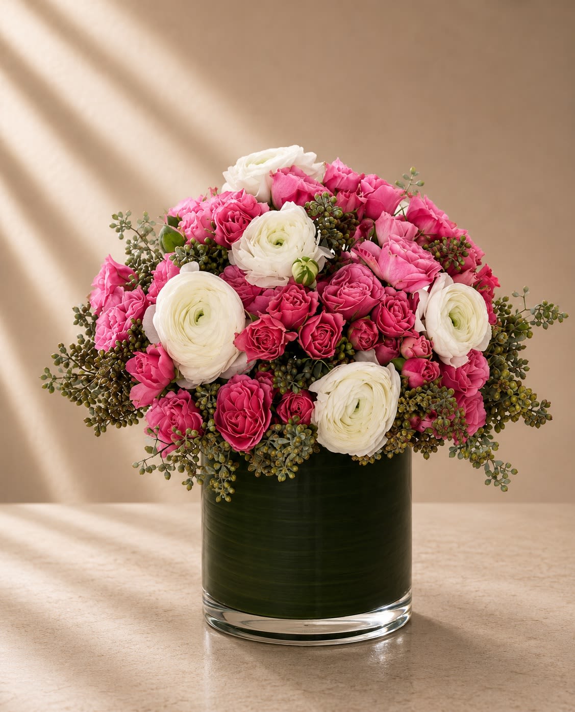Blush Satin Reverie - Soft, feminine, and effortlessly luxurious, Blush Satin Reverie is a romantic couture arrangement designed with delicate textures and graceful movement. Featuring lush white ranunculus paired with abundant hot pink spray roses and airy seeded eucalyptus, this floral composition feels refined, modern, and timelessly elegant.  The layered ranunculus blooms add softness and sophistication with their intricate petal structure, while the vibrant pink roses create richness, romance, and playful charm. Cascading eucalyptus-inspired textures bring natural movement and an elevated garden-style finish, all beautifully presented in a sleek leaf-lined glass cylinder vase.  Designed to feel fresh, romantic, and upscale, this arrangement is perfect for birthdays, luxury gifting, baby showers, anniversaries, bridal celebrations, or elegant everyday décor.