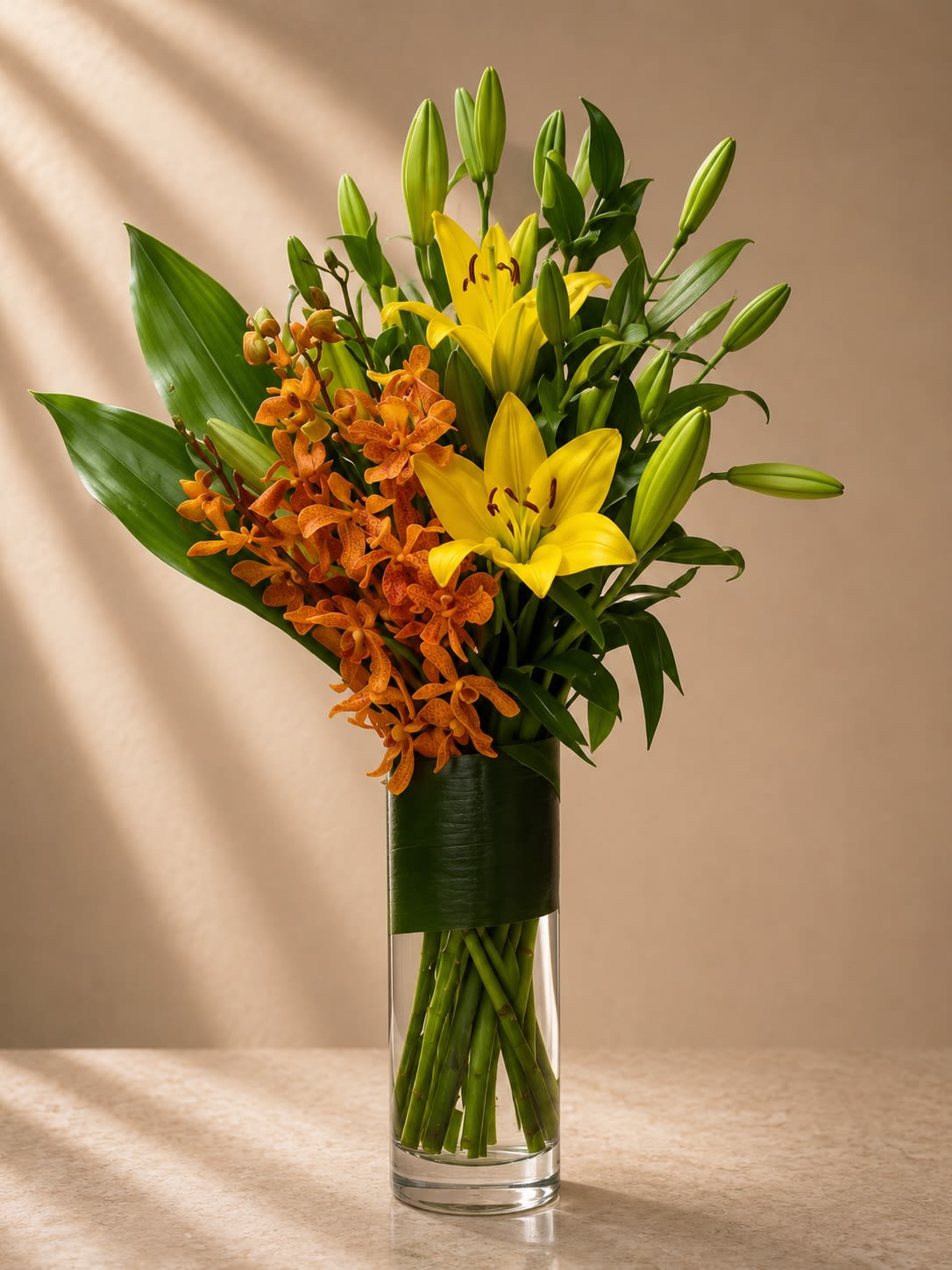 Golden Ember - Bold, radiant, and luxuriously tropical, Golden Ember is a striking vertical arrangement designed with vibrant golden lilies and rich burnt-orange mokara orchids. Styled in a sleek cylinder vase with modern leaf wrapping, this design delivers warmth, movement, and elevated contemporary elegance.  The glowing yellow lilies symbolize joy and prosperity, while the exotic orchids add texture, sophistication, and a couture-inspired tropical flair. The asymmetrical composition gives the arrangement an artistic, gallery-style presence perfect for luxury interiors.  This arrangement feels energetic yet refined — ideal for clients seeking something unique, upscale, and unforgettable.