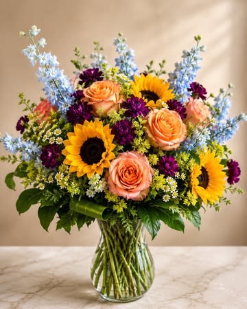 Countryside Charm - Inspired by the beauty of a blooming countryside garden, Countryside Charm is a cheerful arrangement filled with vibrant colors, soft textures, and natural elegance. Bright sunflowers, peach roses, delicate blue delphinium, rich purple blooms, and seasonal accents come together to create a fresh and inviting design that feels as if it were gathered straight from a cottage garden.  The combination of warm golden tones, soft peach hues, cool blue accents, and lush greenery creates a beautiful balance of color and movement. Designed in a clear glass vase, this arrangement brings a touch of rustic charm and timeless beauty to any home or celebration.  Perfect for birthdays, thank-you gifts, get-well wishes, congratulations, housewarmings, or simply brightening someone’s day, Countryside Charm is a joyful expression of nature’s beauty and a wonderful way to make someone feel special.  Substitution Policy  Flower varieties and colors may vary due to seasonal and market availability. If substitutions are necessary, they will be made with flowers of equal or greater value while maintaining the overall style, color palette, size, and beauty of the arrangement.