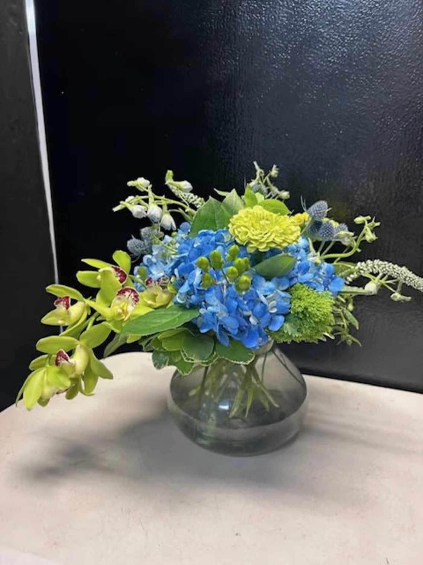 exotic and bold  - Bright blue hydrangeas take center stage in this exotic and bold flower arrangement, paired with lime-green orchids and yellow-green chrysanthemums for a vivid, high-contrast birthday bouquet or congratulatory gift. Smaller white and pale-blue accent blooms fan outward to create a lively, airy silhouette, while broad green leaves anchor the composition so the person you're celebrating feels brightened and noticed.  Arranged in a simple round glass vase, the design reads modern and fresh-perfect for everyday gifting, milestone celebrations, or to say "well done" with personality. The mix of hydrangeas, orchids, chrysanthemums and supporting blooms delivers a mix of texture and color that holds visual interest from every angle; the clear vase keeps care simple and showcases the saturated stems.