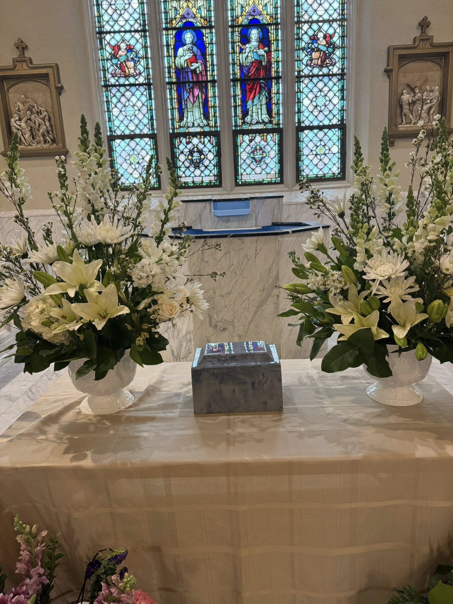 Matching sympathy arrangements in white  - Two matching white sympathy arrangements provide a calm, reverent focal point for a funeral service or memorial. These tall, airy floral displays are designed as paired altar or podium pieces for a ceremonial setting, giving the person you're honoring a sense of dignity and comfort while framing the space with quiet grace.  Each arrangement features white lilies, snapdragons, chrysanthemums and small white rose and other white blooms will   set against soft green foliage, arranged to feel open and uplifting rather than heavy.   Ideal for memorial services or to accompany a casket or urn, these paired designs express sympathy and respect for family and friends gathering to remember someone special. Order guidance and sizing are available at checkout-our team can confirm delivery and placement details to ensure the arrangements arrive ready to frame the service exactly as pictured.