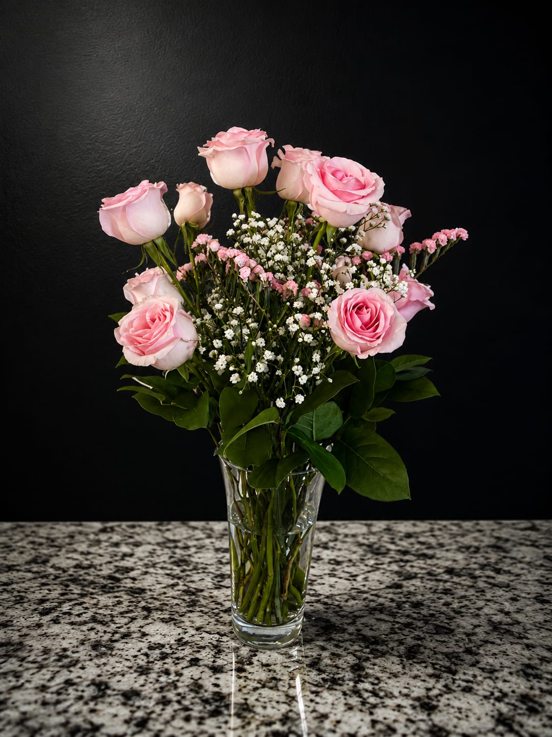Pink Pearl - Pale pink roses with fuller blush centers create an immediate soft-focus impression-perfect for a birthday bouquet, anniversary roses, or a tender "thinking of you" gift. The Pink Pearl flower arrangement reads as romantic and airy, the gentle pinks and crisp whites sending a message of warmth and affection that will make the person you're thinking of feel truly cherished.  Clustered around sprays of tiny white filler flowers and glossy green foliage, the roses sit naturally in a clear glass vase with stems visible through the water for a fresh, garden-picked look. The palette stays light and pastel-pink, white, and green-balanced by the vase's clean lines; overall size and scale suit a tabletop or bedside display without overwhelming a room.  Choose this arrangement when you want to mark an intimate moment or brighten an ordinary day-the kind of bouquet that feels personal and considered. Arrives ready to display in its clear glass vase; keep in fresh water, out of direct heat, and trim stems if needed to extend vase life.