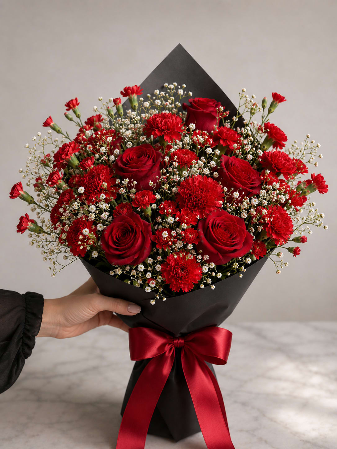 Red Velvet Rose - Deep, sensual red roses and clustered red carnations make an immediate statement in this romantic bouquet-perfect as a Valentine's Day bouquet, an anniversary roses gift, or a heartfelt everyday surprise for the one you love. The dramatic red-and-black palette reads formal and passionate, and the addition of soft white baby's breath gives the arrangement a tender, airy contrast so the recipient feels both cherished and adored.  Tightly hand-tied for a lush, compact silhouette, the arrangement pairs matte black paper and black floral wrap with a wide red satin ribbon for a polished, gift-ready finish. Expect a full composition of deep red roses, red carnations, and delicate white filler that feels rich and celebratory without being fussy.  Give this to a partner, spouse, or special someone when you want them to feel truly seen and treasured-ideal for anniversaries, Valentine's Day, or any romantic milestone. The bouquet arrives dressed and ready to present; trim stems and refresh water if transferring to a vase to keep blooms looking their best.