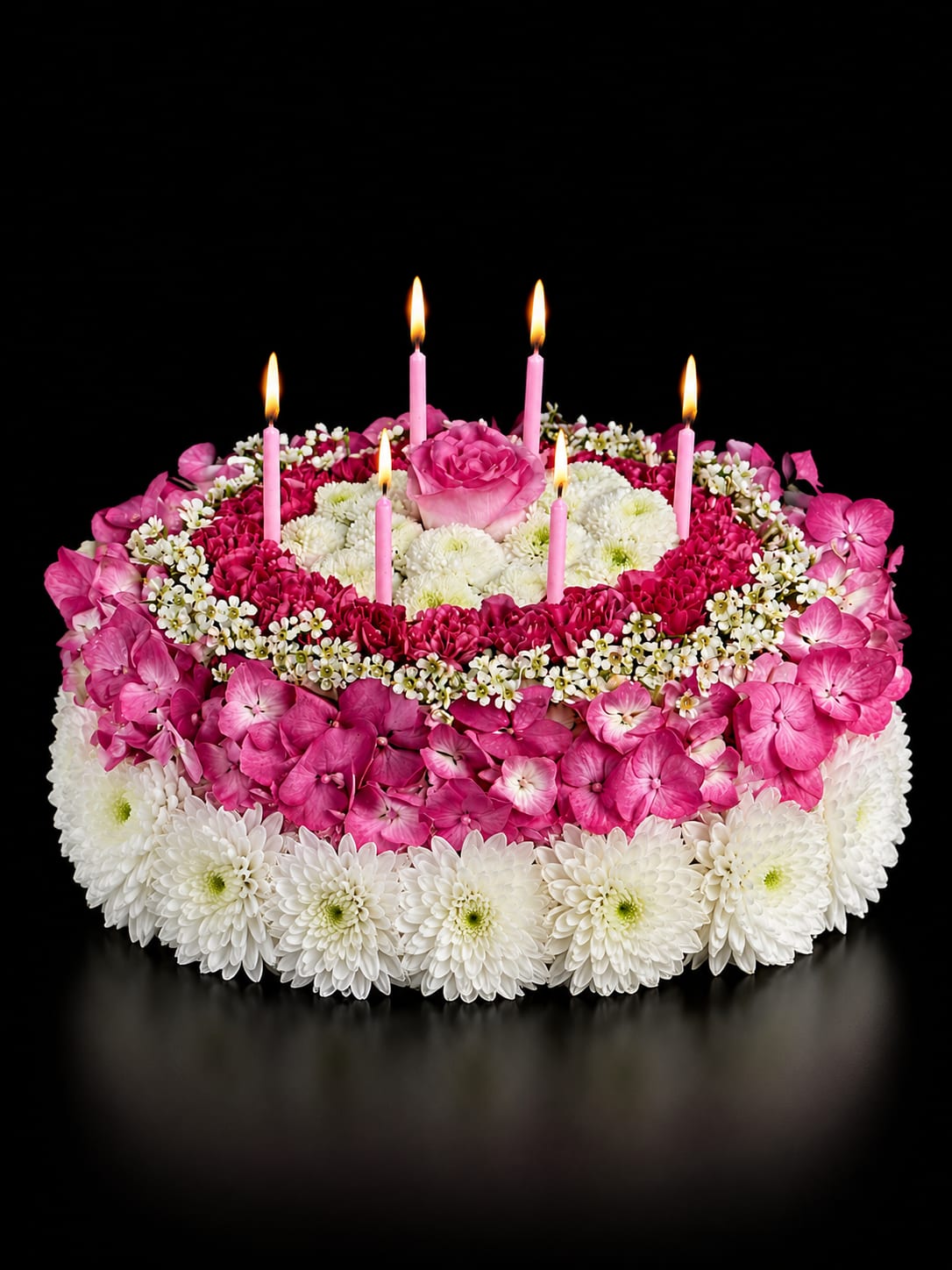 Sweet Celebration - Imagine presenting a floral "cake" that reads like a party in bloom - a Sweet Celebration flower arrangement perfect for a birthday bouquet or any festive milestone. The tiered, round composition looks like a cake from afar, with a single pink rose crowning the top and slim pink taper candles tucked among the blooms so the person you're honoring feels celebrated the moment the door opens.  Close-up, tightly packed bright pink roses and mixed pink florals form the middle band while white chrysanthemum-like blooms create a crisp outer edge and a soft center, giving a contrast of texture and color that pops against darker surroundings. The palette of pink, white, and mixed tones keeps the look cheerful and giftable; the slim pink candles add height and a playful, candlelit mood without overpowering the blossoms.  Made to mark birthdays, congratulations, or any joyful occasion, this arrangement reads as both festive and thoughtful - the kind of bouquet that makes someone feel truly seen on their special day. Treat as you would a fresh bouquet: keep in a cool spot away from direct sun and replace water daily to extend bloom life.