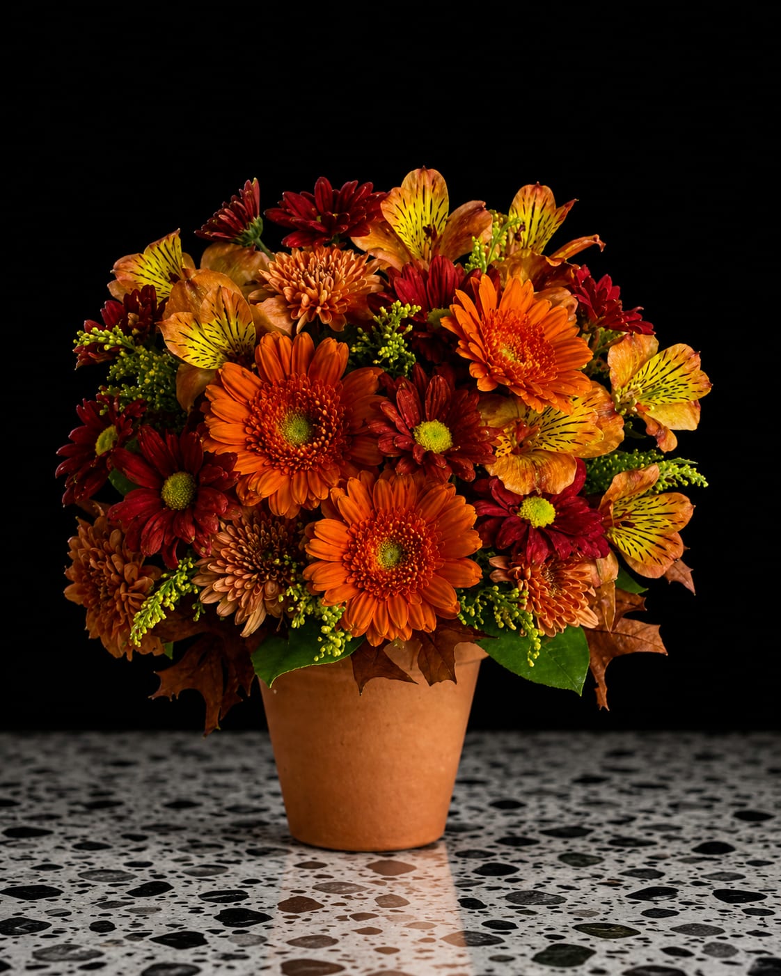 Harvest Colors - A lively harvest of orange, red and golden blooms makes this fall flower arrangement a cheerful choice for seasonal decorating or as a birthday, thank-you, or hostess gift. The low, rounded silhouette of this flower arrangement and the terracotta pot give it a cozy, autumnal feel-perfect for a Thanksgiving centerpiece or to brighten someone's day with warm, harvest colors.  Packed with chrysanthemums, gerbera daisies and speckled alstroemeria, the composition shows off daisy-like shapes and variegated petals for visual interest. Small green filler accents and a few brown leaves add texture and keep the palette grounded, while the simple terracotta pot reinforces the rustic, homey vibe. Sized to sit easily on a table or mantle, it's an everyday seasonal arrangement that will make the person you're thinking of feel celebrated and seen.