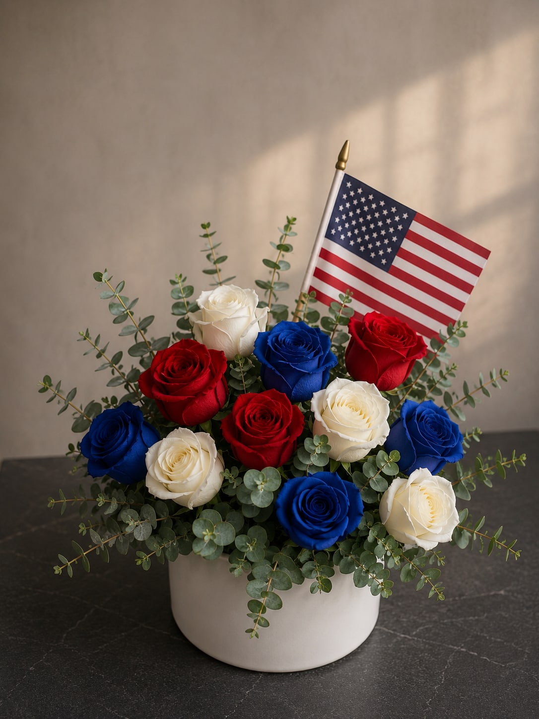 Spirit of freedom - A low, proud cluster of red, white, and blue roses creates an instantly patriotic statement-perfect as a Fourth of July centerpiece or a congratulatory arrangement for a veteran or graduate. The mix of saturated red and blue roses against crisp white blooms reads bright and celebratory, while the American flag rising behind the bouquet makes the sentiment unmistakable: a thoughtful way to honor service, celebrate a national holiday, or mark a milestone with pride.  The roses are arranged full and rounded in a clean white ceramic vase, accented with airy eucalyptus branches that add soft green texture and a gentle, fragrant lift. The compact, low profile makes it ideal for tabletops, mantels, or as a dignified display at a reception. Order with confidence for delivery to homes or events-this arrangement is designed to feel festive on arrival and to convey respect and celebration to the person you're honoring.
