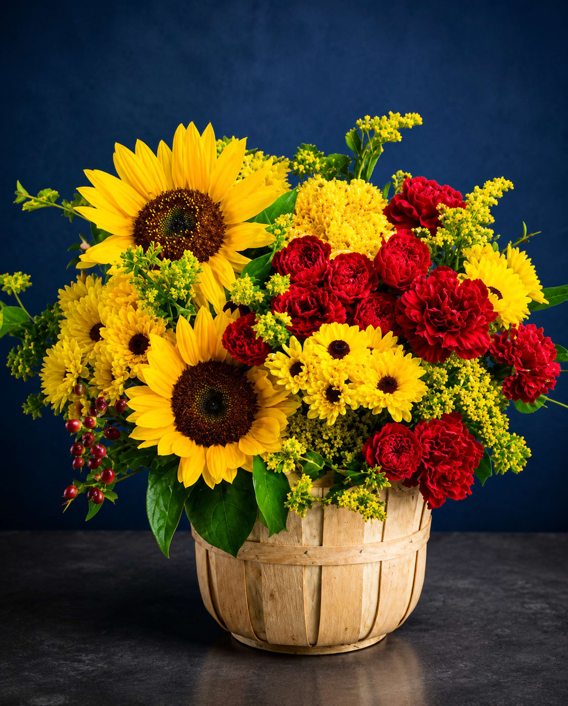 Golden Harvest - Large, golden sunflowers take center stage in this autumn-ready flower arrangement, a cheerful gift-perfect bouquet for birthdays, congratulations, or a warm-season thank-you. The bright yellow blooms paired with clusters of red carnations and yellow chrysanthemums create a bold, celebratory look that will make the person you're thinking of feel genuinely seen and celebrated.  Arranged in a rustic wooden basket, the design reads full and abundant: large sunflowers, mixed-color chrysanthemums, and rich red carnations are accented by small filler blooms and berries for texture and a garden-fresh feel. The warm yellow, red, and green palette pops against darker backgrounds, making it a standout centerpiece or entryway accent.  Ideal for sending seasonal cheer or marking a special milestone, this arrangement arrives ready to display in its basket - no vase required. Its sturdy construction and lively color story make it an effortless, heartwarming present that brightens a room and lifts spirits.