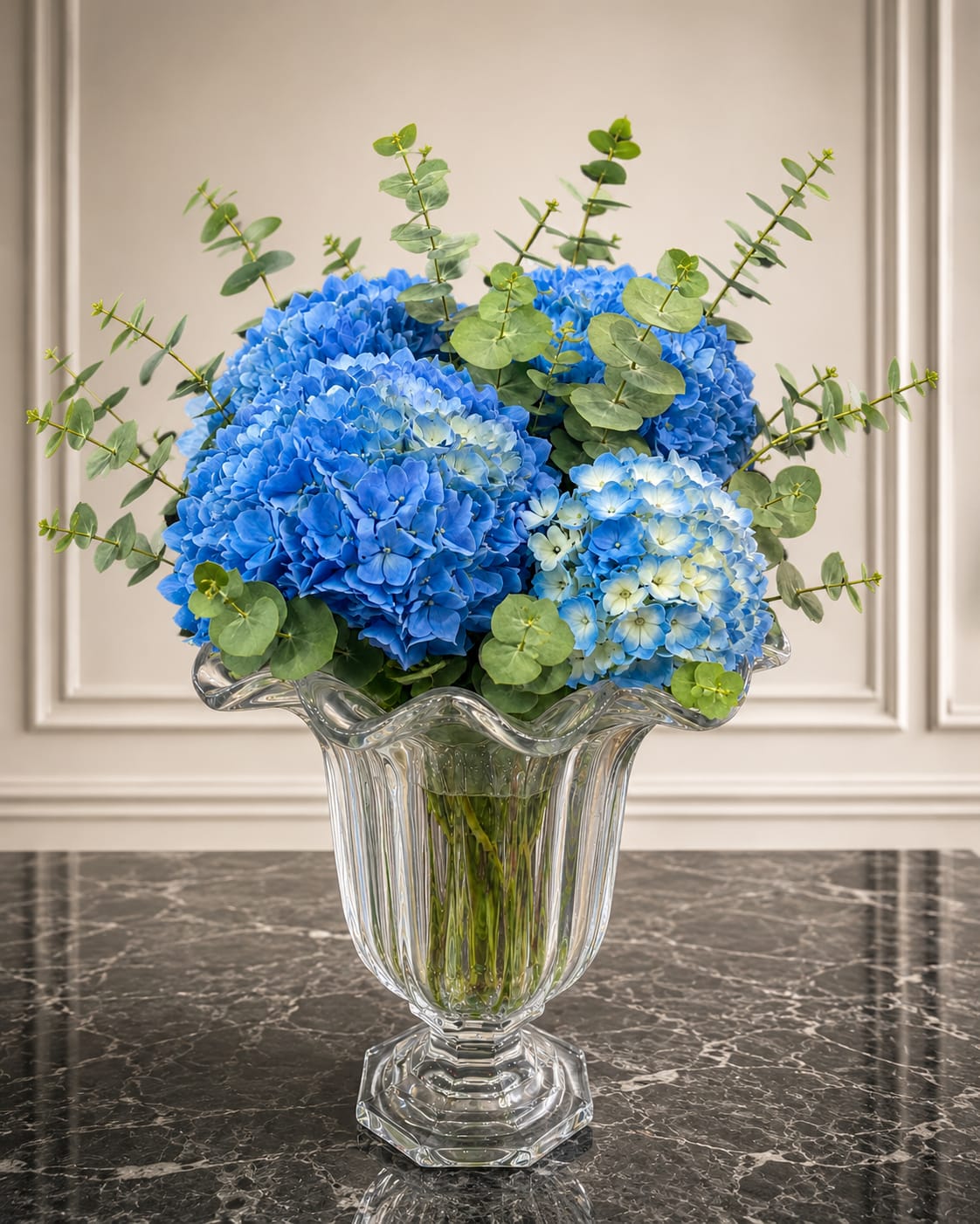 Blue Serenity - Rounded clusters of blue hydrangeas-from vivid cobalt to pale sky blue-create an immediate sense of calm and refreshment, making this flower arrangement a perfect birthday bouquet, sympathy arrangement, or everyday gift to soothe and uplift. Tall eucalyptus stems rise above the blooms, adding airy texture and a soft green contrast so the person you're thinking of will feel seen and comforted by the design's peaceful presence.  Set in a clear fluted glass vase that reveals the stems and lends a polished, formal touch, the arrangement pairs cool blues, fresh greens, and crisp white accents for a serene palette that fits living rooms, bedside tables, or office desks. The open silhouette and rounded hydrangea heads make a generous visual statement while remaining easy to place-order with confidence for celebrations, get-well wishes, or as a thoughtful way to say you care.