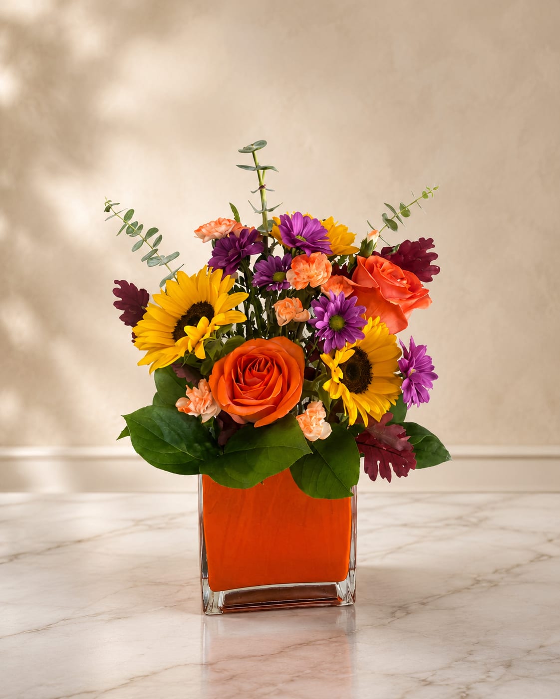 Autumn Radiance - Bright orange roses and sunny yellow gerbera and sunflower heads make this autumn-inspired flower arrangement a burst of cheer - perfect as a birthday bouquet, congratulations gift, or just-because surprise to brighten someone's day. The vivid orange, purple, yellow, and pink palette reads bold and joyful; the person you're thinking of will feel noticed and celebrated the moment this arrives.  Compact and textured, the design pairs bright roses with yellow gerbera daisies, small purple daisy-like blooms, peach carnations and pops of dark burgundy foliage for contrast. Eucalyptus-like stems lift the composition slightly above a full, round center, all presented in a clear square glass vase filled with orange decorative filler that reinforces the warm color story.  Choose this arrangement when you want a lively, upbeat floral gift that reads festive without being fussy - ideal for friends, coworkers, or anyone who loves vibrant color. The compact vase makes it easy to place on a desk or table; fresh care is standard for mixed bouquets (keep water topped up and out of direct heat) to keep blooms looking their best.