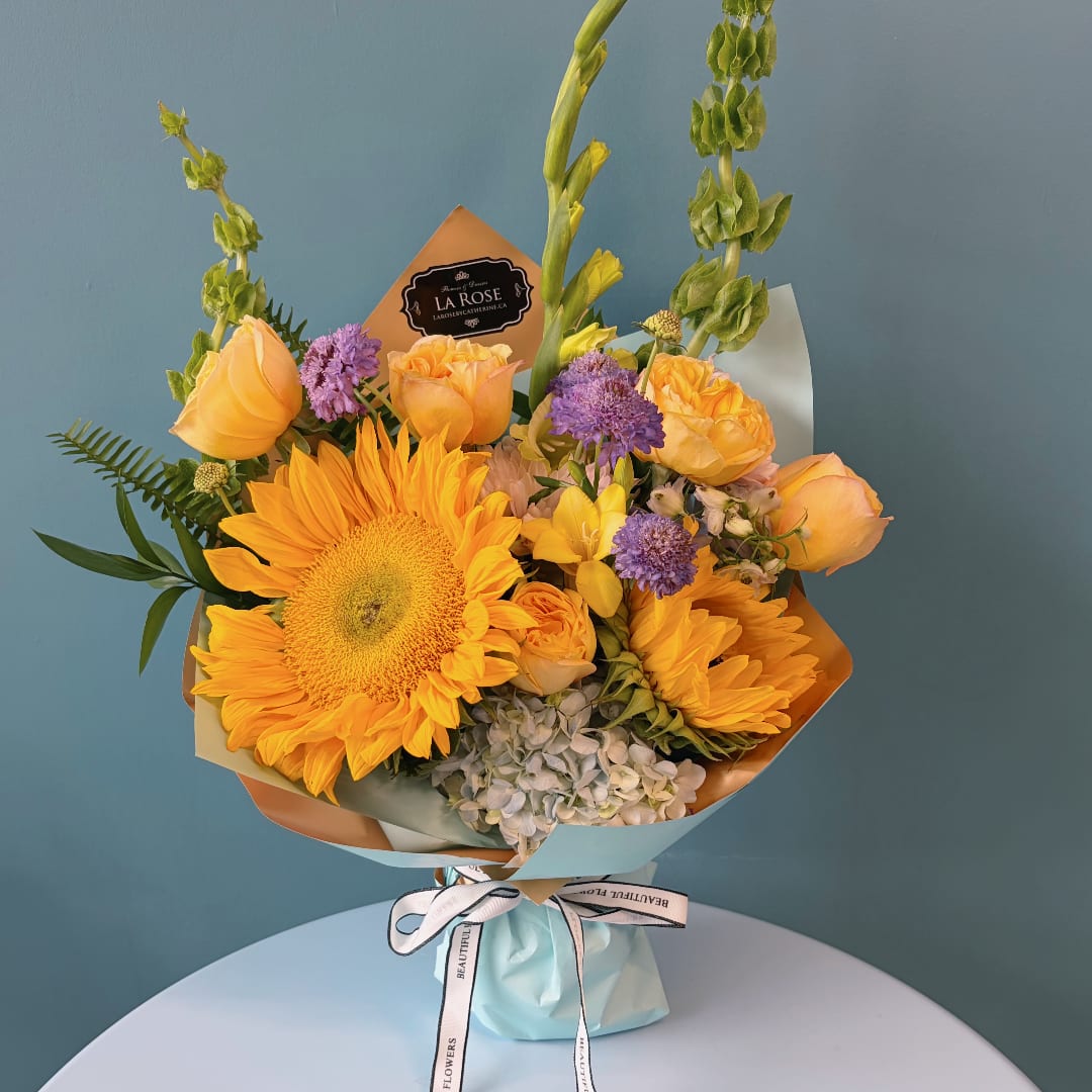 Bouquet of yellow sunflowers, peach roses, and purple flowers wrapped in pastel paper