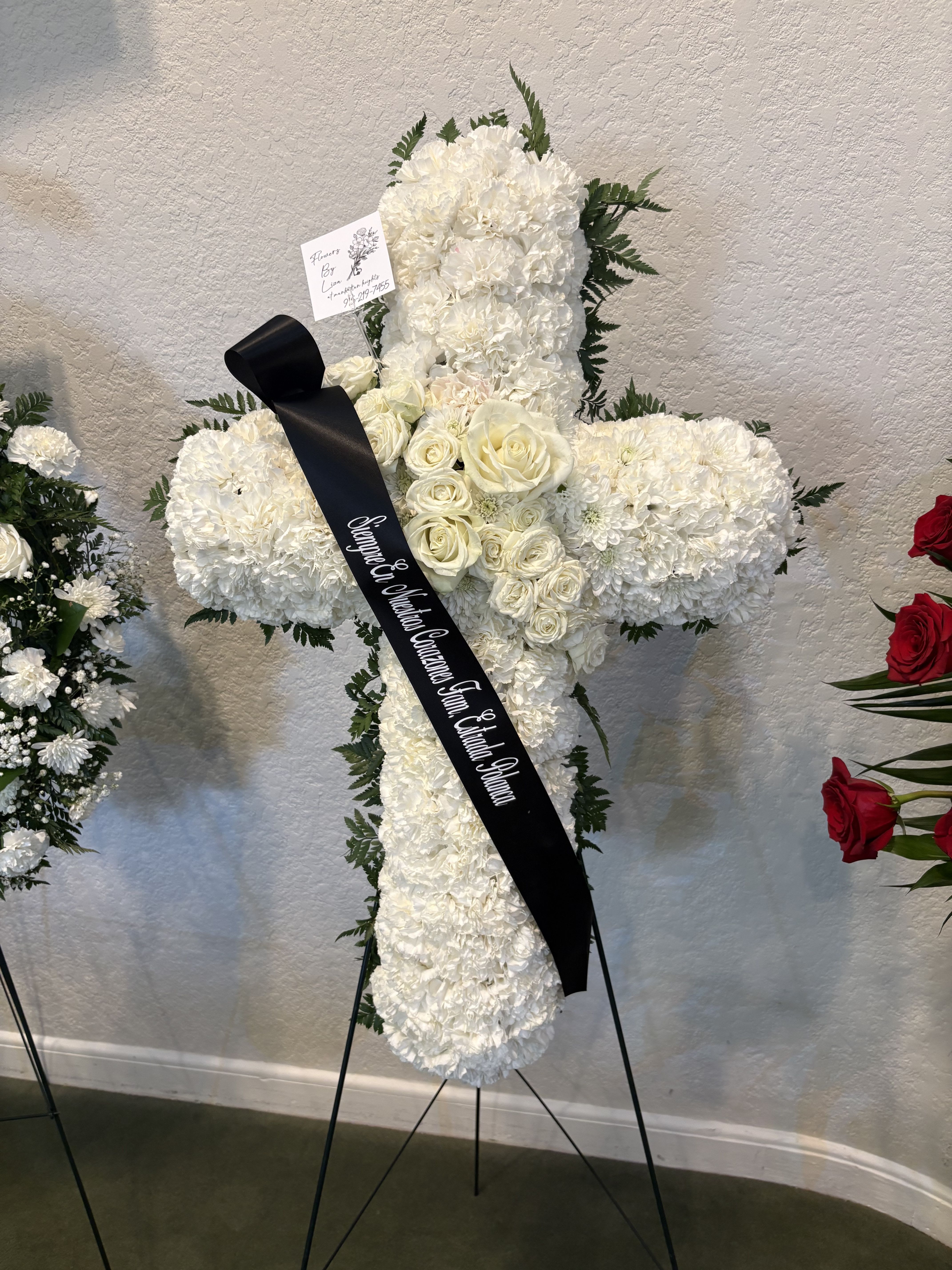 White sympathy cross - A solemn standing sympathy cross that offers quiet, traditional reverence for a funeral or memorial service. This white sympathy cross frames white roses, carnations, and chrysanthemums into a dense, symmetrical silhouette so the person you're honoring feels remembered with dignity and care.  Clusters of white roses gather near the center while fuller white carnations and chrysanthemums form the arms and vertical sections, their soft textures contrasted by deep green foliage around the edges. A black memorial ribbon is draped diagonally across the face and the arrangement is finished with a sturdy metal easel and a card for your message, giving it a formal, respectful presentation at a service.  Appropriate for graveside, chapel, or memorial displays, this cross conveys sympathy and respect without being ornate-a meaningful way to say you're thinking of them. Standard sizing places it among large standing tributes; best kept out of direct heat and misted lightly if the service is outdoors to maintain freshness.
