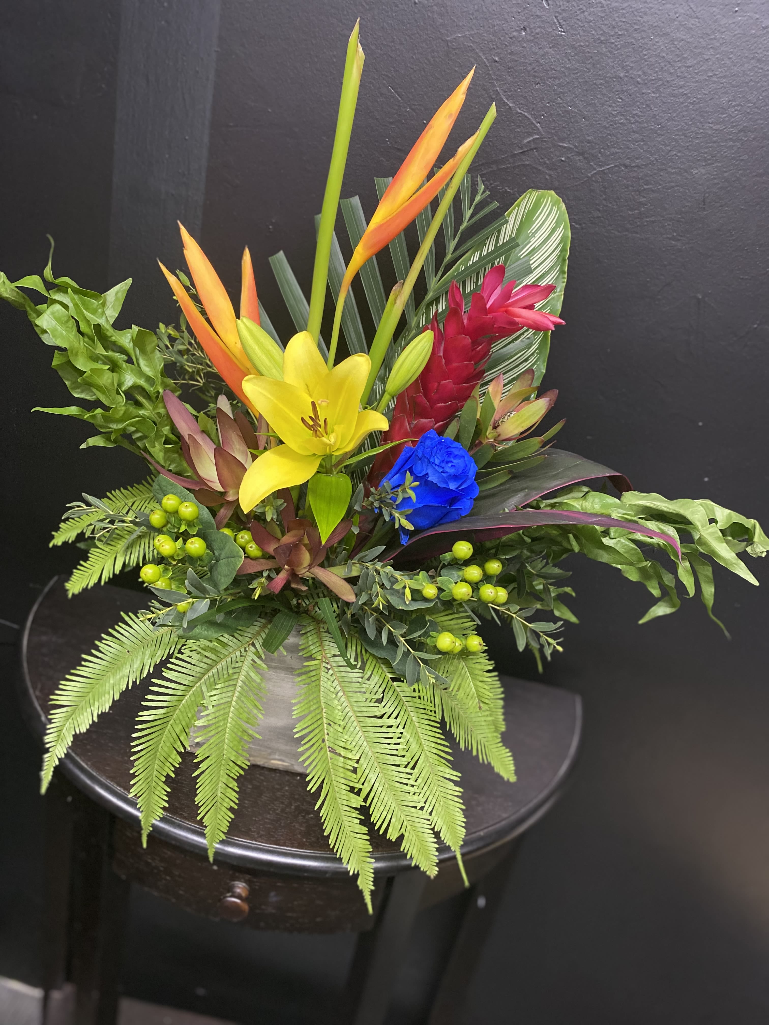 Tropical Oasis - A bright yellow lily and vivid blue rose anchor this tropical bouquet, making a bold statement perfect for a birthday bouquet or any celebratory gifting moment. The combination of orange bird-of-paradise and a striking red ginger flower gives the arrangement an exotic, festive energy that will make the person you're thinking of feel celebrated and noticed.  Layered with broad and feathery tropical foliage and small green berry accents, the design reads dense and textured-highly saturated colors that pop against a darker display setting. Presented in a ceramic vase, this mixed-color arrangement blends lilies, roses, and classic tropical blooms for long-lasting visual impact; its dramatic palette is ideal for party centerpieces, congratulatory deliveries, or a standout reception display.