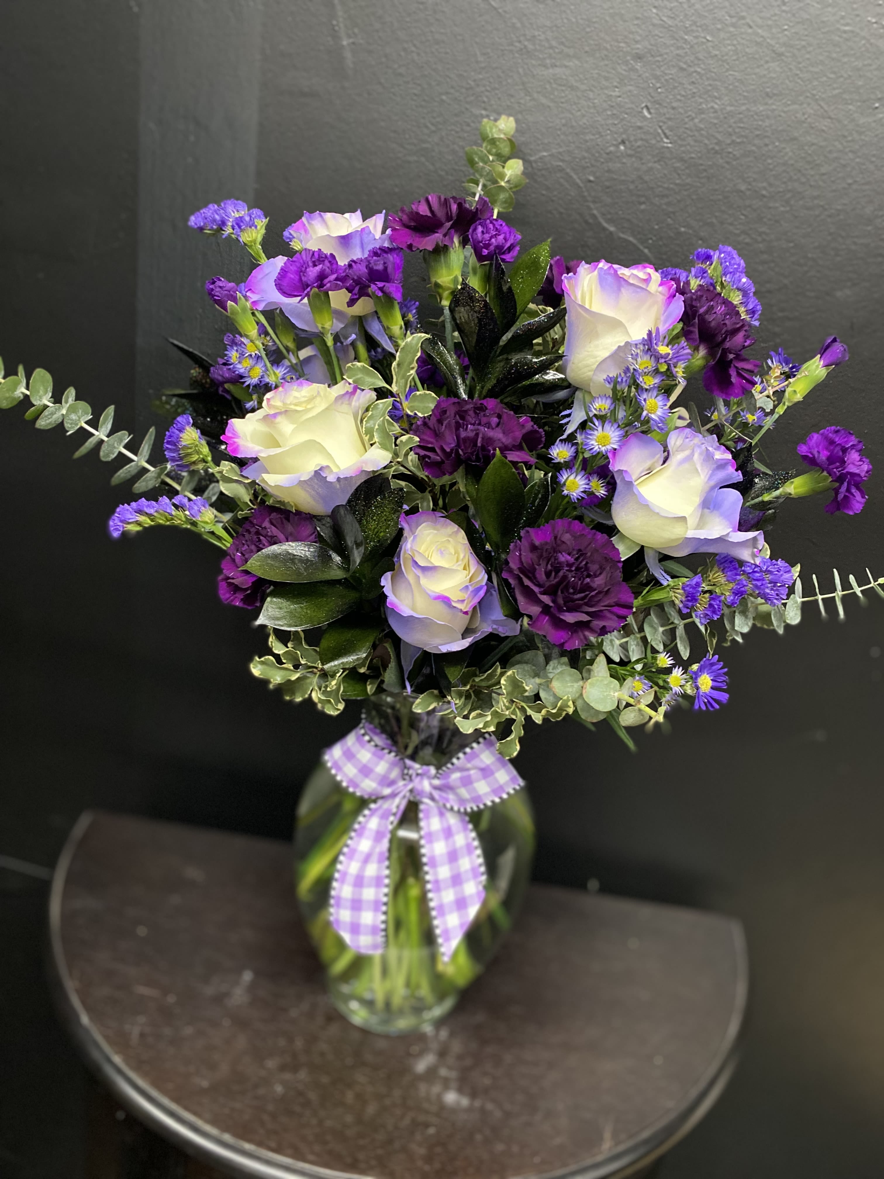 Purple Prairie Delight - White roses edged in lavender meet deep purple carnations in this cheerful spring bouquet-perfect as a birthday bouquet or a bright-thinking "just because" arrangement. The mix of purple, white, and fresh green creates a lifted, celebratory look that will make the person you're thinking of feel noticed and celebrated, whether you're marking a milestone or simply sending a warm hello.  The arrangement pairs fragrant white roses with lavender edges, rich purple carnations, and small purple aster-style filler blooms for a lively mix of shapes and textures. Eucalyptus and glossy greenery frame the rounded, full bouquet, adding soft scent and contrast. It arrives in a clear glass vase finished with a purple gingham ribbon for an easy, gift-ready presentation.  This arrangement suits springtime celebrations, birthdays, or a thoughtful surprise to brighten someone's day. Available in a single vase size as shown; to keep blooms fresh, change the water every two days and recut stems at an angle when refreshing the vase.