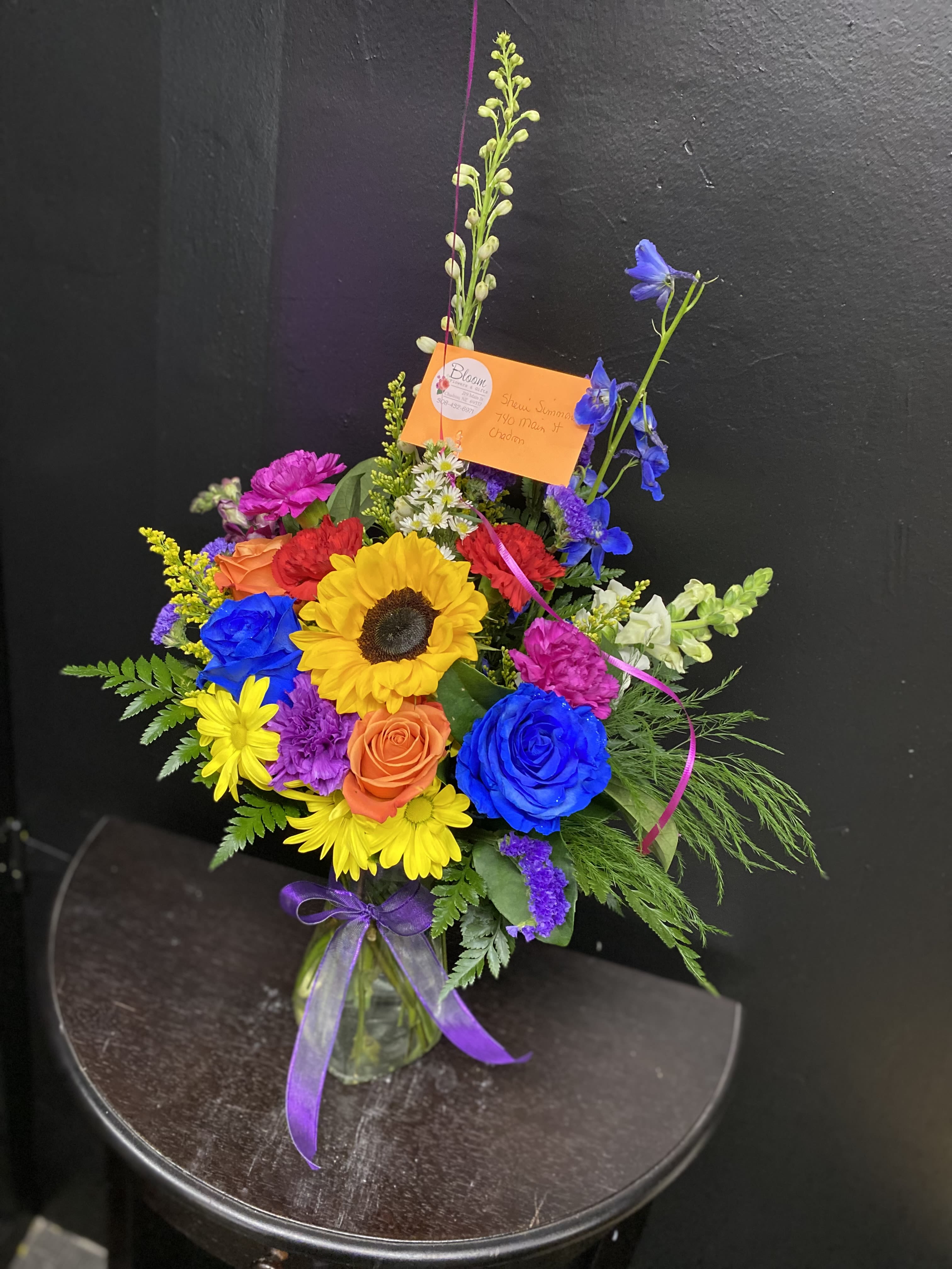Bright Side Blooms - A bold sunflower takes center stage in this Bright Side Blooms flower arrangement, a cheerful mixed bouquet perfect for birthdays, congratulations, or a thank-you gift. The bright yellow sunflower paired with vivid blue and orange roses creates an uplifting focal point that will make the person you're thinking of feel noticed and celebrated.  Pink and red carnations, small daisy-like asters, and tall snapdragon spikes add playful texture and vertical movement, while layered greenery gives the design a full, garden-style silhouette. Delivered in a clear glass vase and finished with a purple ribbon and an orange note card, the palette of yellow, blue, and mixed tones reads joyful and festive without feeling fussy.  Choose this arrangement when you want someone to smile-whether marking a milestone or just brightening their day. The size and height from the snapdragons make it stand out on a table or mantel; change the message on the included orange card to tailor the sentiment, and enjoy easy care by keeping the vase filled with fresh water.