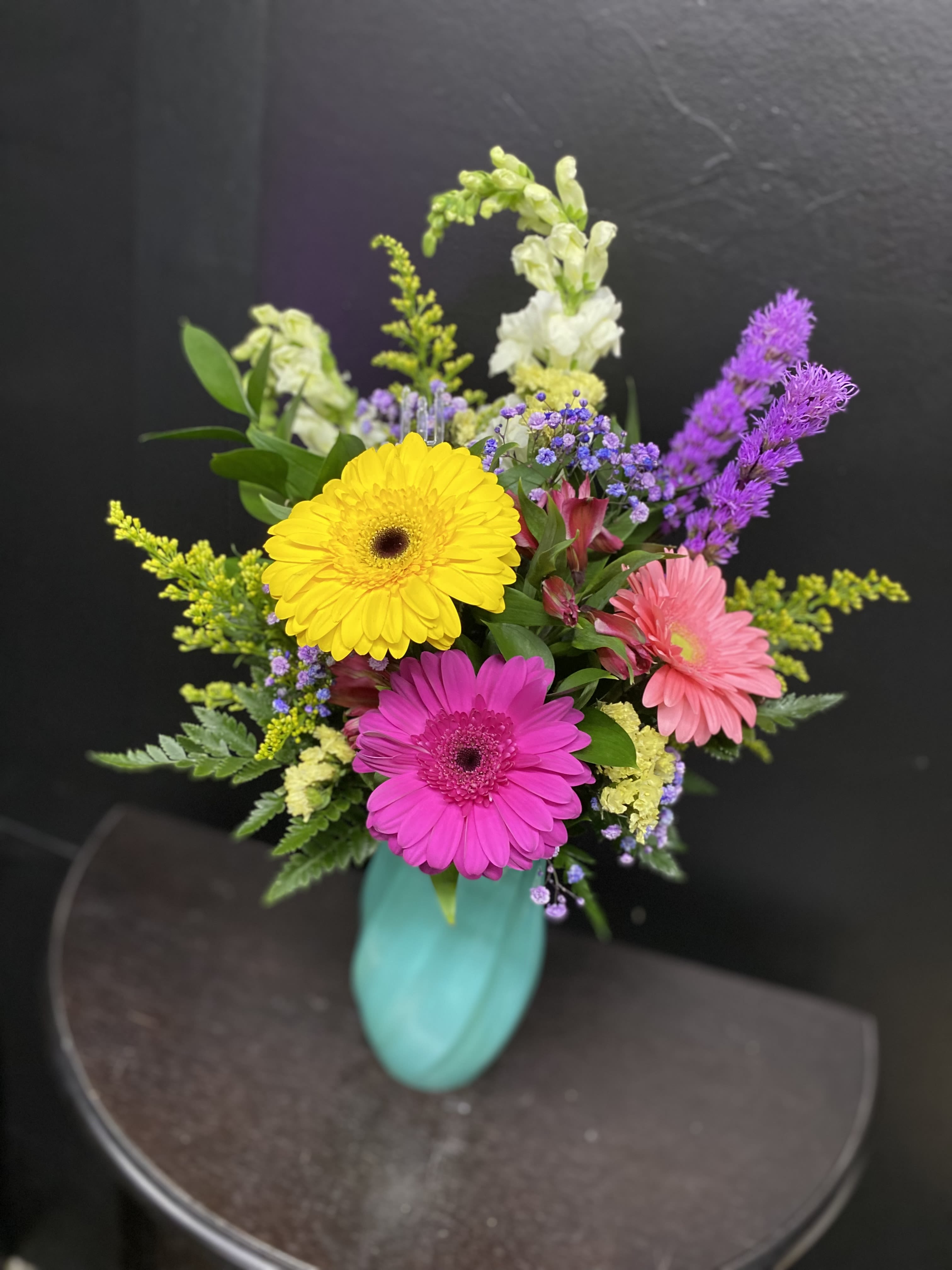 Wildflower Jubilee - Three bold gerbera daisies-bright yellow, magenta, and soft pink-create an immediate burst of joy in this birthday bouquet, perfect for birthdays, congratulations, or any celebratory moment. Tall white stock adds vertical drama while airy purple and yellow fillers give the arrangement a garden-fresh texture; the overall effect is lively and contemporary, designed to make the person you're thinking of feel noticed and celebrated.  Arranged in a striking turquoise vase that pops against the stems, the bouquet balances vibrant color with a clean, upright silhouette. Flowers include gerbera daisies, white stock, asters and other seasonal fillers in mixed pink, yellow and purple tones. The layered height from the stock and the round gerbera heads creates visual interest from every angle, making it an excellent centerpiece or bedside pick-me-up.  This arrangement suits recipients who appreciate cheerful, modern florals-friends, coworkers, or family marking a happy occasion. Vase included; for best longevity, refresh water every two days and trim stems slightly when you change the water to keep blooms at their peak.
