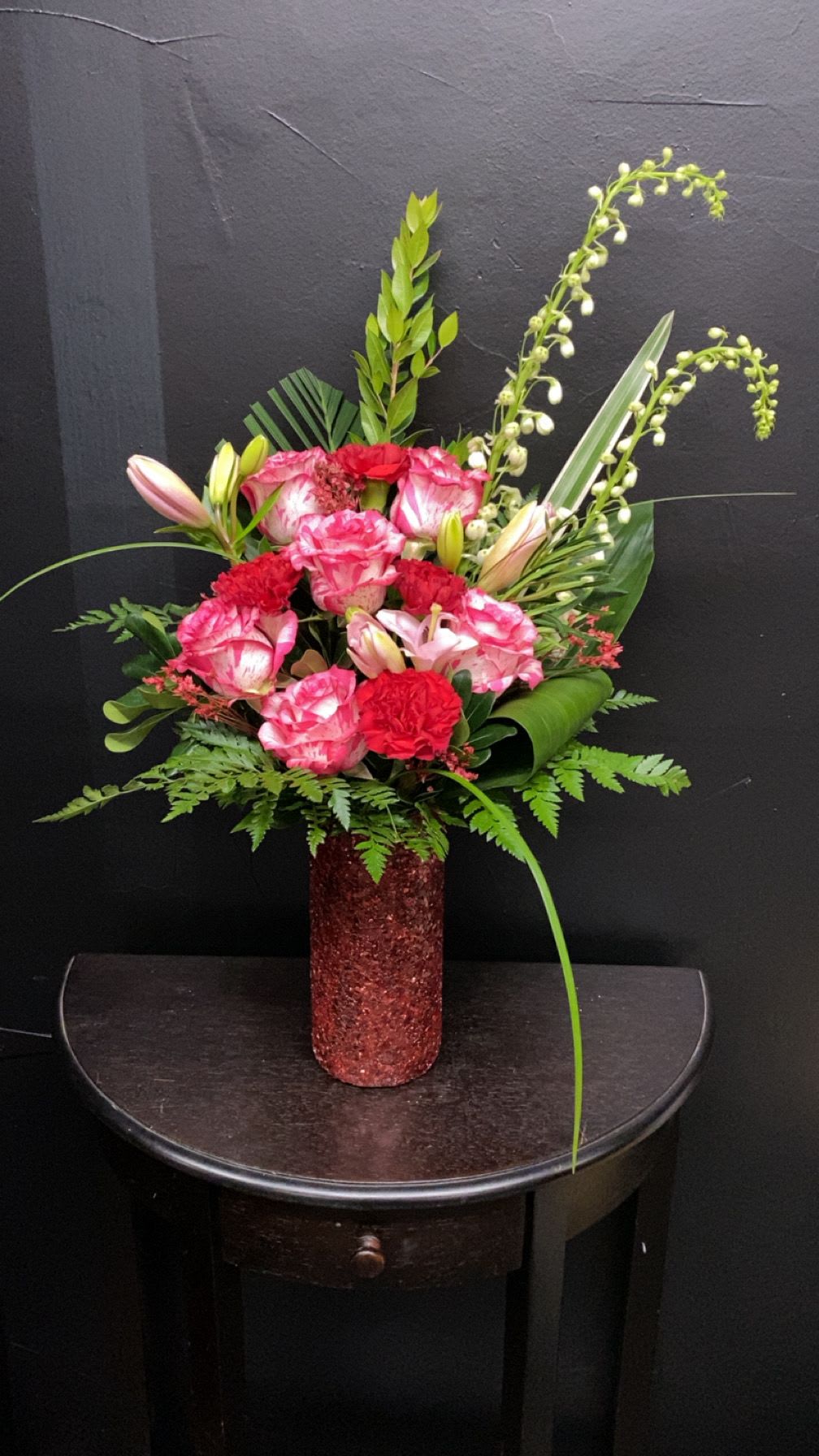 Crimson Charm - A bold, celebratory bouquet designed to make someone feel deeply noticed - this upright floral arrangement blends pink-and-white blooms with vivid red carnations for a romantic birthday or anniversary bouquet that still reads as joyfully festive. The dramatic, tropical-like silhouette created by tall, arching stems and layered greenery gives the recipient an immediate sense of occasion, while the glittery red cylindrical vase turns the whole gift into a keepsake-ready display.  The mix includes pale lily buds peeking through a dense cluster of pink-and-white flowers alongside bright red carnations, creating contrast in both color and texture - soft petals against structured buds and glossy foliage. Finished in a sparkling red vase, the arrangement stands tall and eye-catching on a dining table, mantel, or entryway; its vertical shape lends presence without sprawling. This is a confident, decorative piece that reads romantic and celebratory at once.  Perfect for marking anniversaries, birthdays, or a seasonal romantic surprise, it's the kind of bouquet that makes the person you're thinking of feel truly celebrated. No special care is needed beyond fresh water every two days and keeping the display out of direct heat to prolong the life of the lilies and carnations.