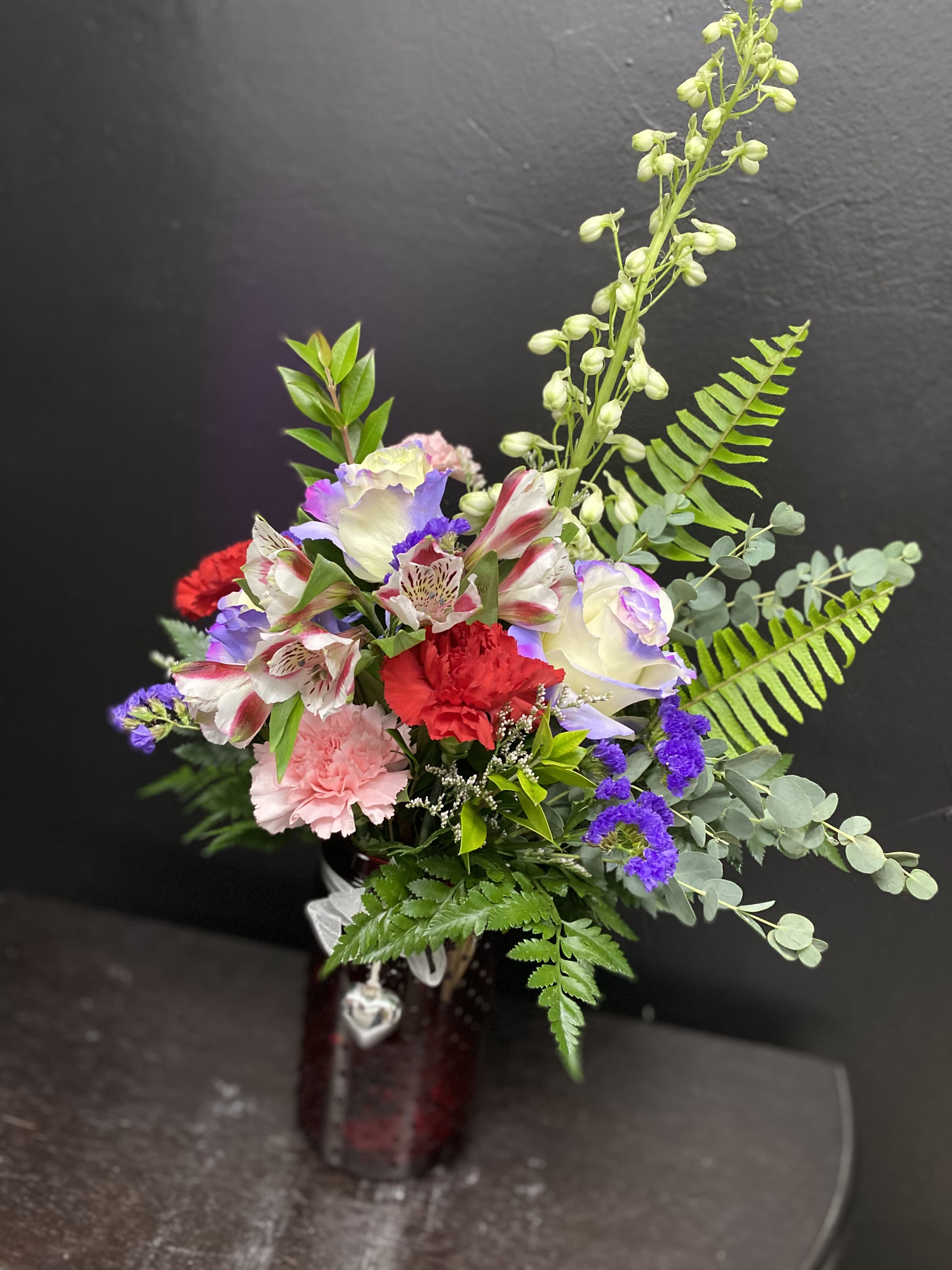 Meadow Melody - A colorful, romantic flower arrangement built to make someone feel truly seen - Meadow Melody is a mixed-color bouquet perfect for anniversary roses, a birthday bouquet, or a "thinking of you" gift. The combination of pink and red carnations with white and blush roses and striped alstroemeria creates a soft, layered look that reads both joyful and intimate, while purple accent blooms add unexpected depth.  This arrangement is hand-arranged with tall fern fronds and eucalyptus-like foliage that lift the silhouette into a loose, airy shape. It arrives in a dark glass vase finished with a decorative ribbon and a small hanging heart charm, giving it a polished, gift-ready presentation. Ideal for celebrating love, friendship, or special milestones - the person you're sending it to will feel cherished and noticed. Care: keep vase water fresh and in a cool spot away from direct sunlight to extend bloom life.