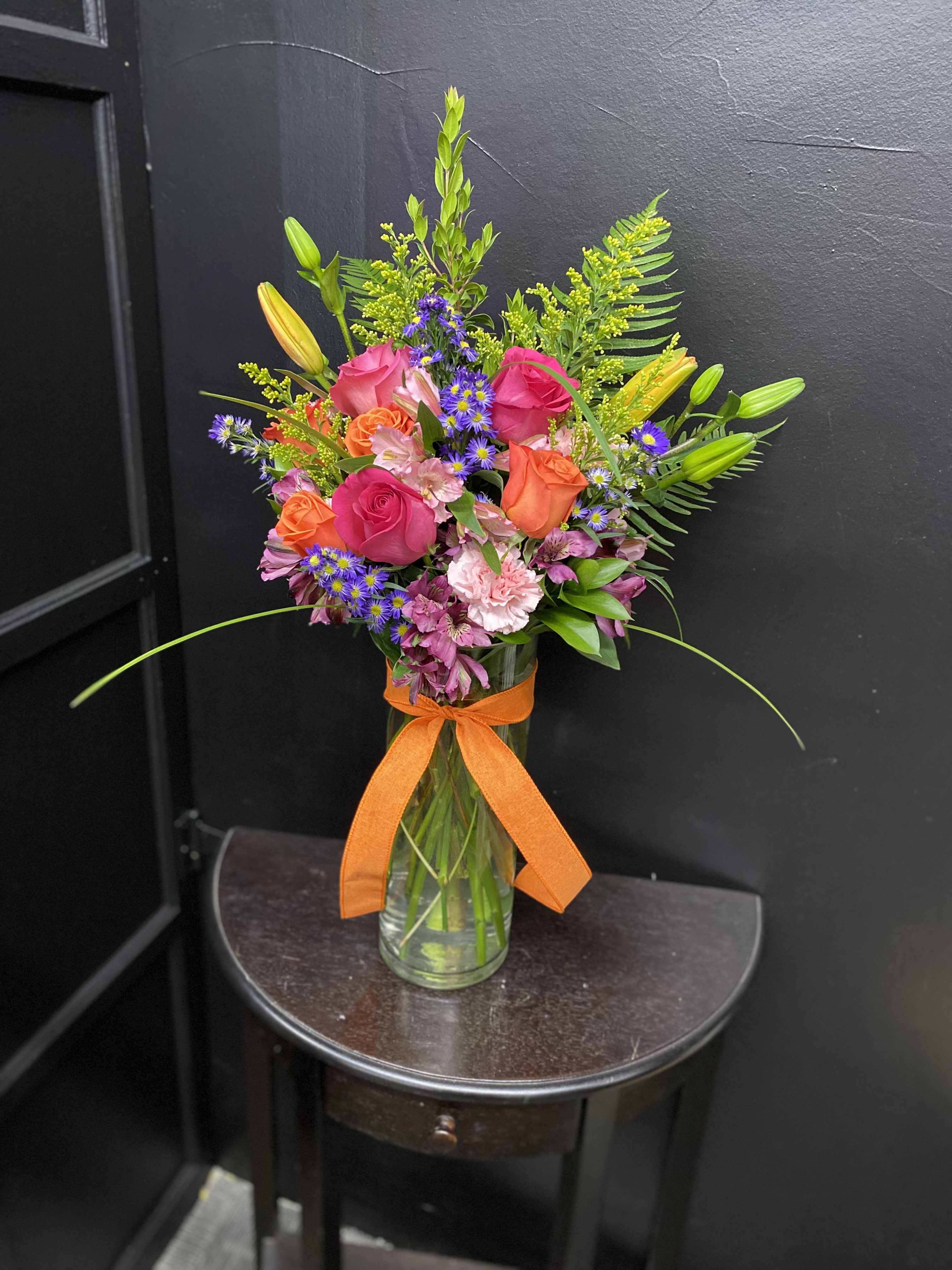 Sunset Celebration - A bright, garden-style bouquet that feels like a warm evening glow-perfect as a birthday bouquet or a congratulations arrangement. Sunset Celebration blends pink and orange roses with yellow lily buds and purple daisy-like asters to create an uplifting color story that reads joyful and celebratory for the person you're thinking of.  This full, upright arrangement pairs soft pink carnations, alstroemeria, and snapdragons with long green accents and airy branching elements for texture and movement. Presented in a clear glass vase finished with an orange ribbon, it has a polished yet casual look that brightens a room. Ideal for spring gifting or any happy milestone, the mix of roses, lilies, asters, carnations and snapdragons is designed to feel fresh and abundant; no special care is needed beyond fresh water and a cool spot away from direct heat to keep blooms looking their best.
