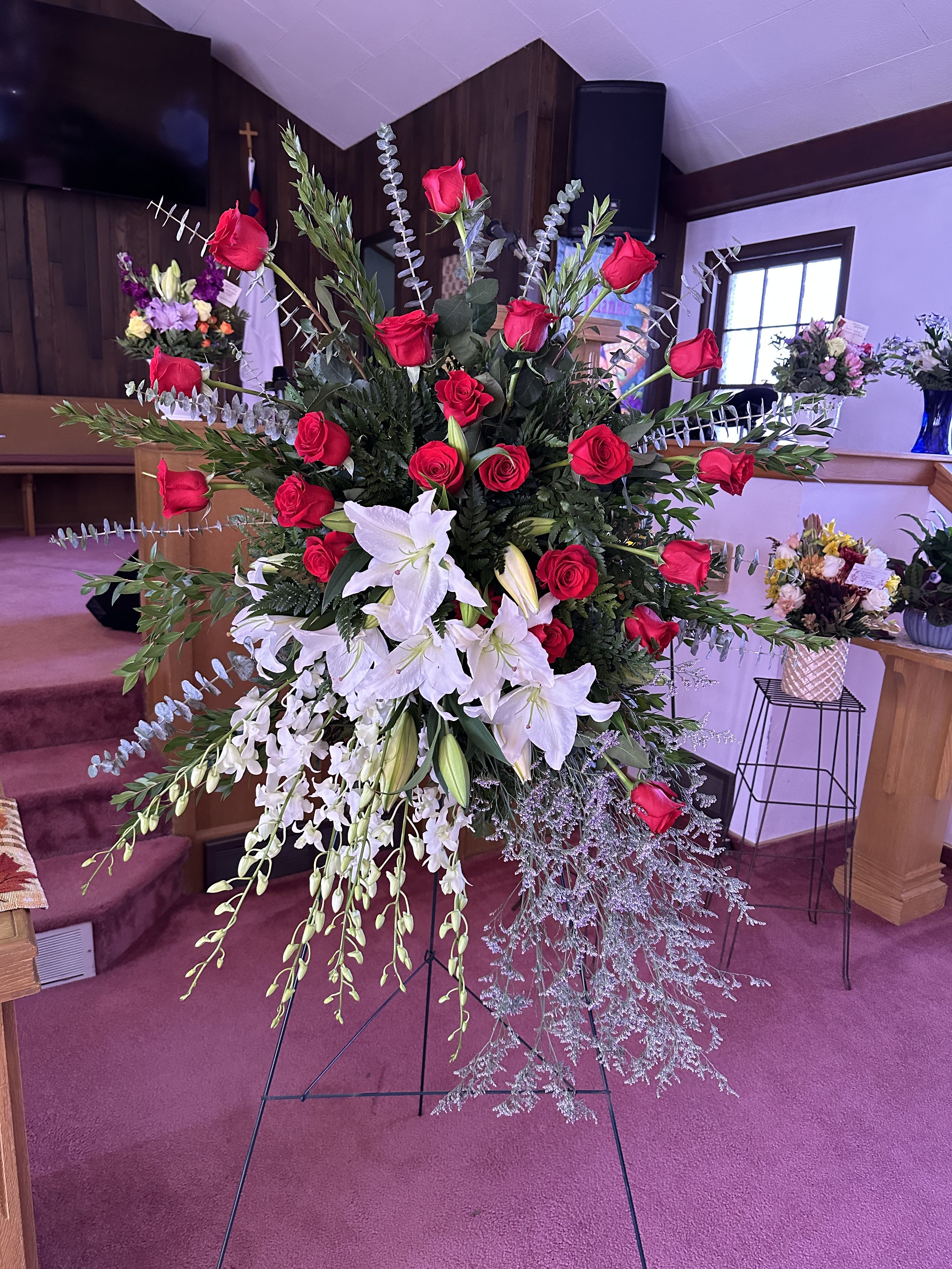 A Life Well Loved - Vivid red roses clustered against a deep bed of greenery create an immediate, dignified presence in this large standing spray-designed as a sympathy arrangement or funeral tribute to honor a life well loved. White lilies near the center lend a serene focal point while touches of purple orchids and silvery, airy foliage give the piece a gentle lift, creating a fan-like silhouette that reads both formal and heartfelt when displayed on the included metal easel stand.  This arrangement combines traditional sympathy flowers-roses, lilies, orchids-with a rich palette of red, white, green, and purple for contrast and depth. Tall vertical height and long, outward-reaching stems provide strong visual impact suitable for church or memorial service displays. The overall effect is solemn yet comforting: the person you're honoring will be remembered with warmth and grace. Available as a large standing spray mounted on a sturdy metal easel; designed for immediate display and arranged to maintain formality throughout the service.