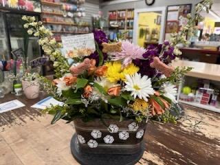 Pet Memorials  - A lush, rounded memorial arrangement that brings warmth and color to a pet remembrance or sympathy setting - this pet memorial flower arrangement blends chrysanthemums, carnations, roses and mixed seasonal blooms to create a comforting display. The mixed color palette of purples, pinks, whites, with touches of yellow and peach offers a gentle, hopeful mood that suits a memorial service, a graveside tribute, or a quiet at-home remembrance for the person grieving a beloved pet.  The design is dense and layered, with large daisy-like chrysanthemums and smaller clustered blossoms set among fresh greenery for texture and depth. Roses and carnations add soft petals and lasting form while the handcrafted dark rectangular planter, finished with white floral accents, makes the piece feel like a thoughtful keepsake. The arrangement sits low and compact on a table or memorial surface, presenting an abundant, cheerful composition without overwhelming the space.  Intended to comfort the person you're thinking of, this arrangement reads as a sincere way to say you remember and care. It's ready to place on a tabletop or memorial site; water and basic care for cut stems will extend the bloom life so the colors remain bright during the days of remembrance.