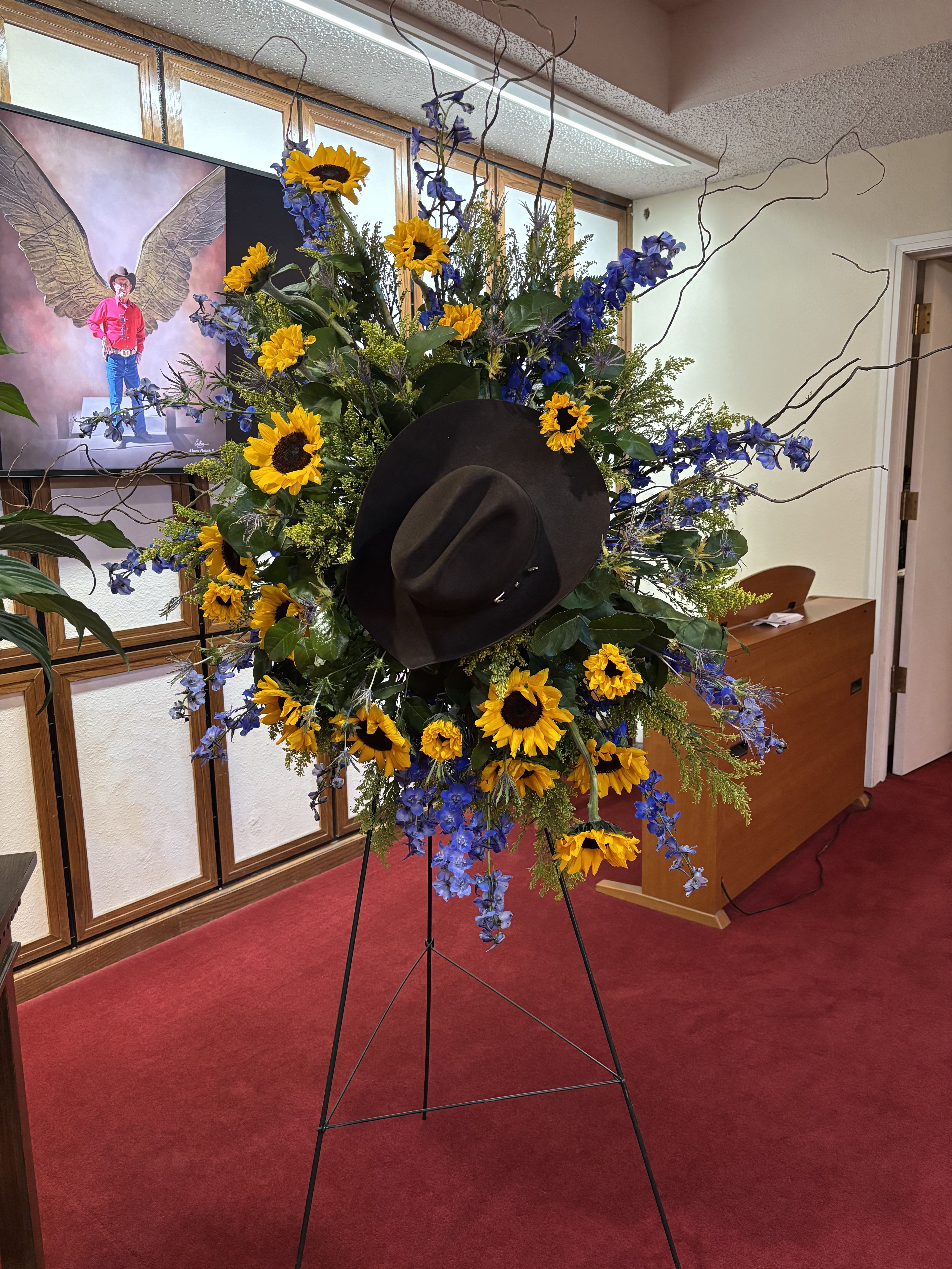 A Cowboy’s Blue Sky - A standing spray with flowers as yellow as the sun and as blue as the sky to remember that special person.  Includes: Easel, Floral Cage, Sunflowers, Salal, Solidago, Curly Willow, and Delphinium 