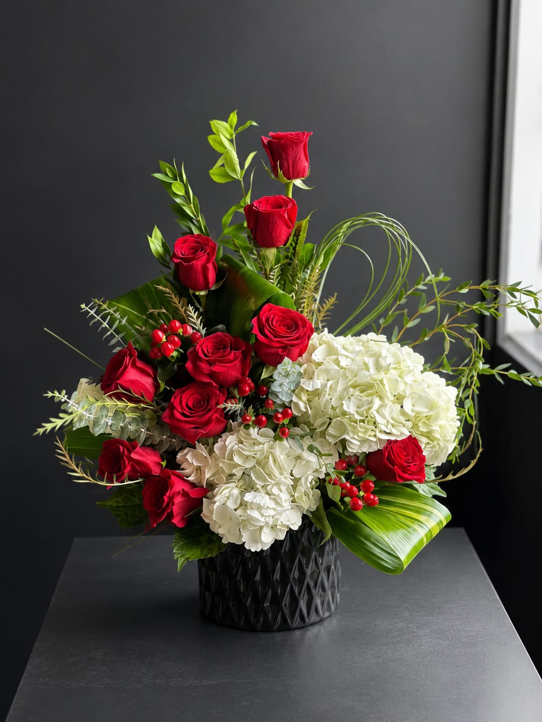 Scarlet Elegance - A timeless arrangement that blends classic romance with modern sophistication. Rich red roses are paired with lush white hydrangeas, vibrant hypericum berries, and premium greenery in a sleek black vase. Designed with graceful lines and contemporary accents, Scarlet Elegance makes a stunning gift for anniversaries, birthdays, sympathy, or simply to express love and appreciation