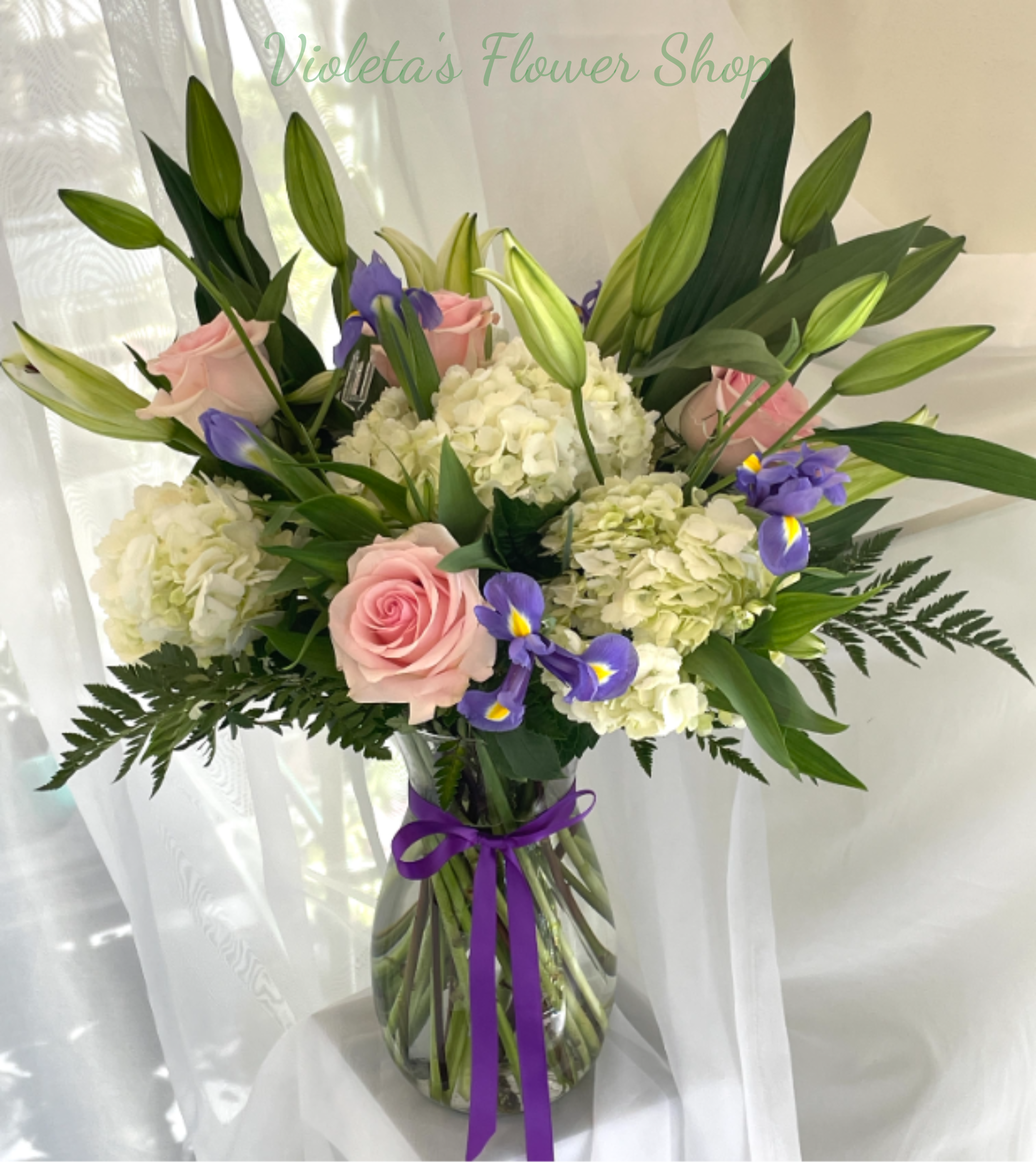 You Are In Our Thoughts. - The arrangement includes roses, hydrangeas, irises, and several unopened lily buds for added height and graceful structure, all arranged in a clear glass vase finished with a purple ribbon for a polished, formal look. The palette of white, pink, purple, and green balances softness and contrast, while the unopened lilies add sculptural lines that will open over time for continued presence.  Send this to someone who needs your sympathy, support, or warm remembrance-the kind of bouquet that helps a person feel seen and cared for. Arrives ready to display in the included clear vase; keep in a cool spot away from direct sun and change the water every two days for the best longevity.