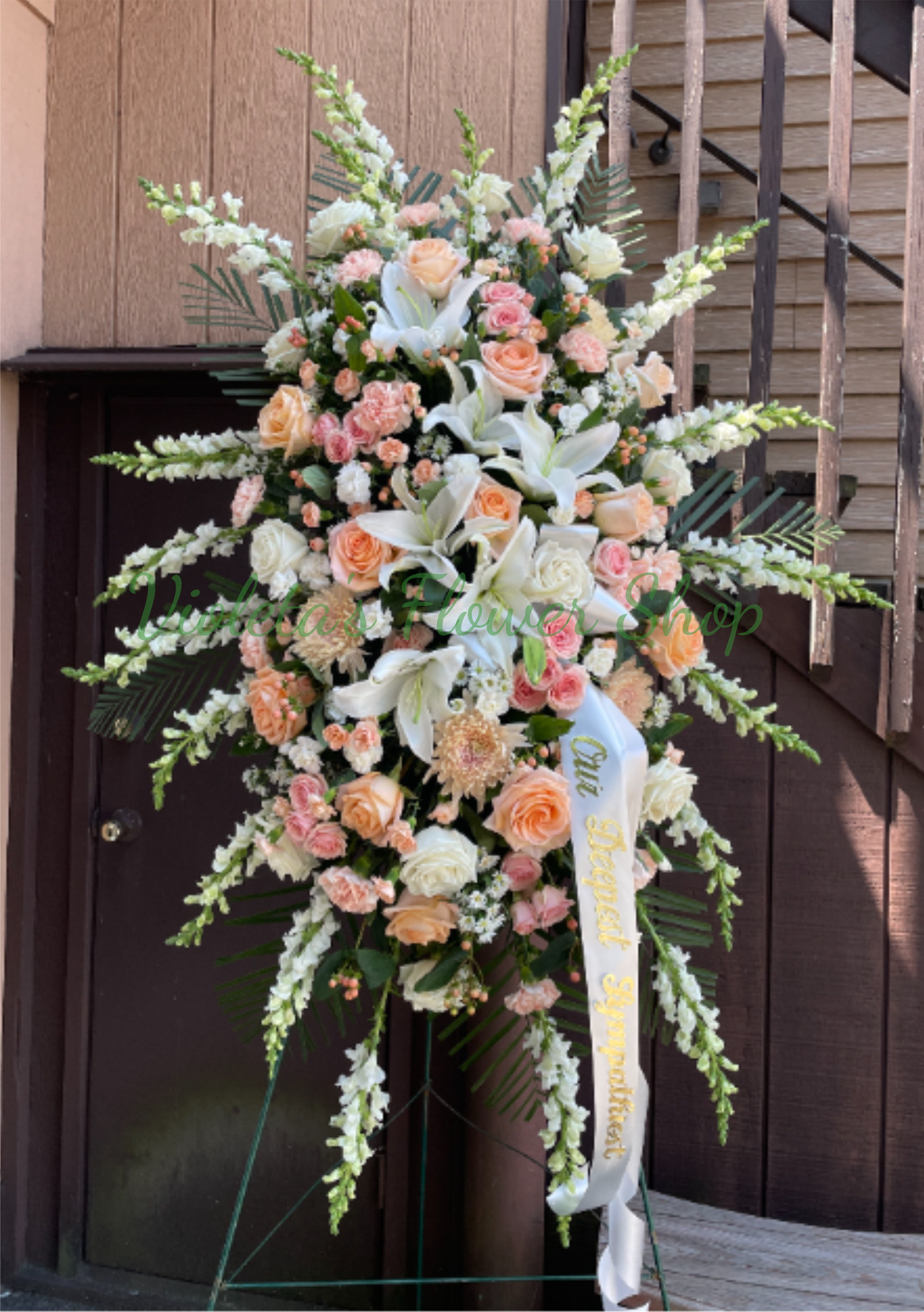Holding You In My Heart Standing Spray - A tall, graceful standing spray that offers a gentle, refined tribute - perfect for sympathy or a memorial service. The arrangement's formal silhouette features a dense center of white lilies and soft peach and blush roses, with long white snapdragon spikes lifting the design into an elegant vertical statement that reads both dignified and comforting to the person you're remembering.  Close-view details include clusters of carnations, chrysanthemums and daisies woven among the roses and lilies for soft texture and lasting depth, all set against fresh greenery. The palette of white, pink and peach creates a warm, peaceful impression; the spray arrives mounted on a dark floral easel with a crisp white ribbon for a respectful presentation. 