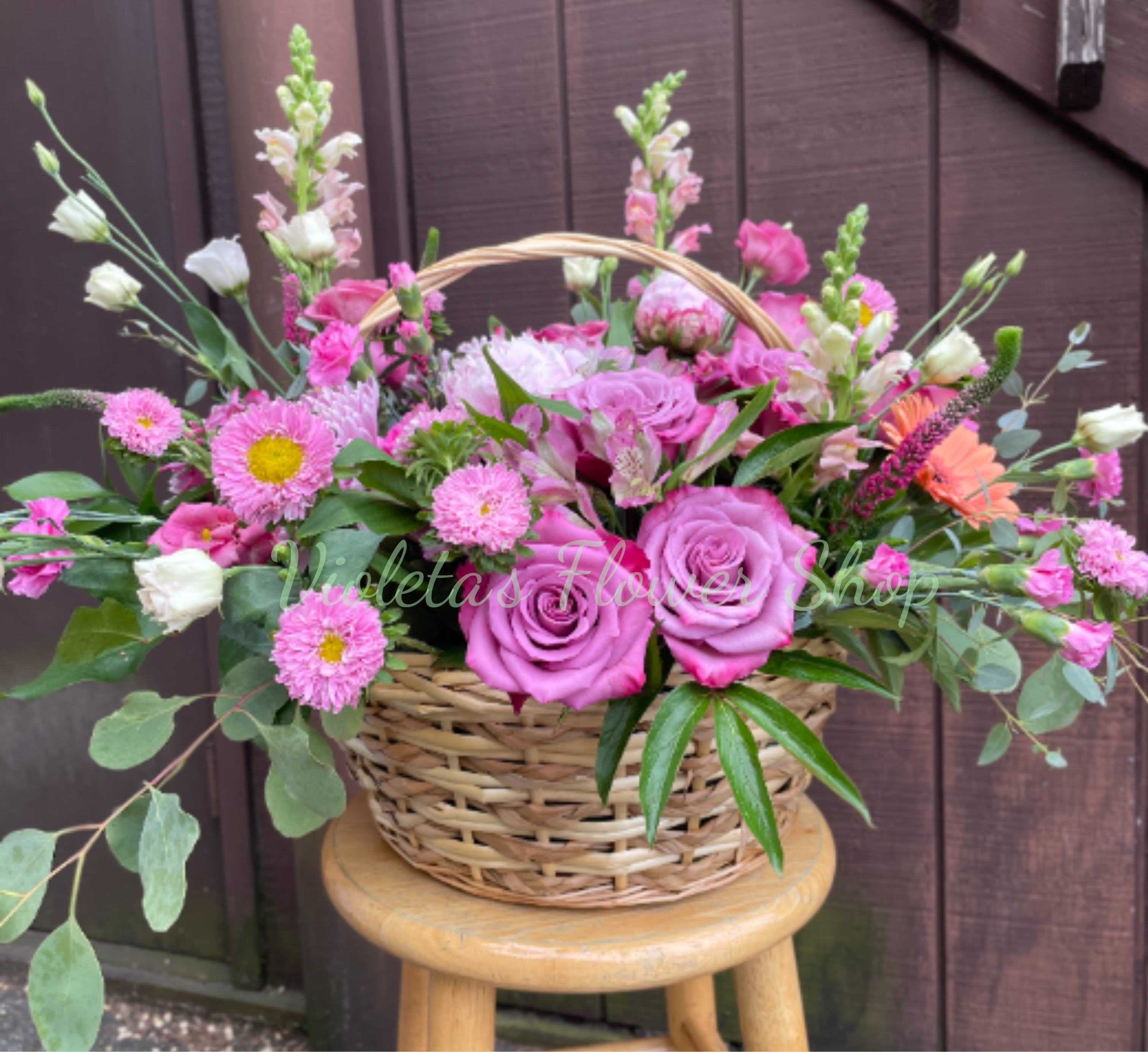 Basket of Pink Blooms - A soft, garden-style basket arrangement that feels like a warm spring morning-perfect as a birthday bouquet, a thank-you gift, or a cheerful surprise for someone you care about. Woven basket and curved handle give this flower arrangement a rustic, hand-tended look while a romantic mix of pink and lavender roses, pale lisianthus, and daisy-like blooms delivers a gentle, uplifting impression the recipient will notice first.  The design layers large lavender-pink roses at the center with airy snapdragons, alstroemeria, lisianthus, and smaller daisy-style flowers fanning outward for a loose, natural silhouette. Fresh green foliage and peach accents add contrast and movement, creating a balanced palette of pink, lavender, green, and touches of peach. The basket keeps everything casual and gift-ready, sized to sit on a table or countertop without overwhelming the room. 
