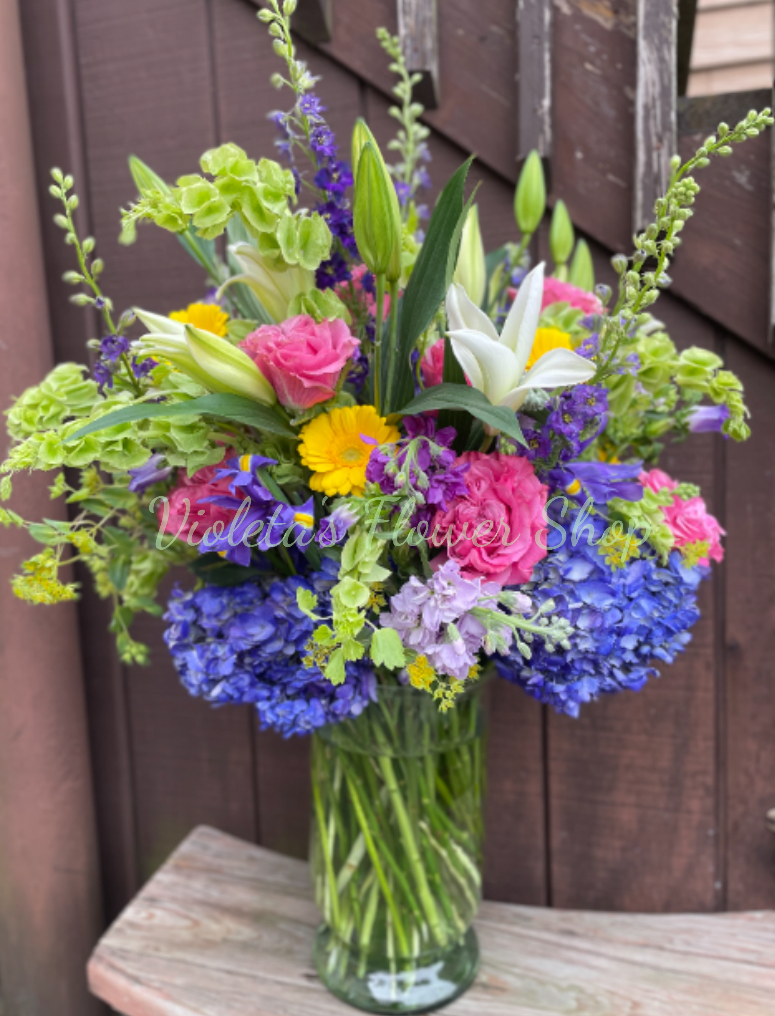 Celebrating You Arrangement - The centerpiece sits in a simple clear glass vase that showcases the layered stems and keeps the look fresh and modern. Dense blue and green blossoms form a rounded base, with taller stems rising above for a full, garden-style silhouette; textures range from soft hydrangea heads to the crisp edges of gerbera-style daisies and the elegant trumpet of lilies. Color notes: bright pink, white, blue, yellow, purple, and green for a lively, versatile palette.  Ideal for welcoming a new baby, saying thank you, congratulating someone on a milestone, or brightening a new home or retiring colleague's day-she'll feel truly celebrated when this arrives. Vase-ready and arranged for visual fullness, this bouquet is low-maintenance: fresh water in the glass vase and a quick trim of stems every few days will help keep blooms looking their best.