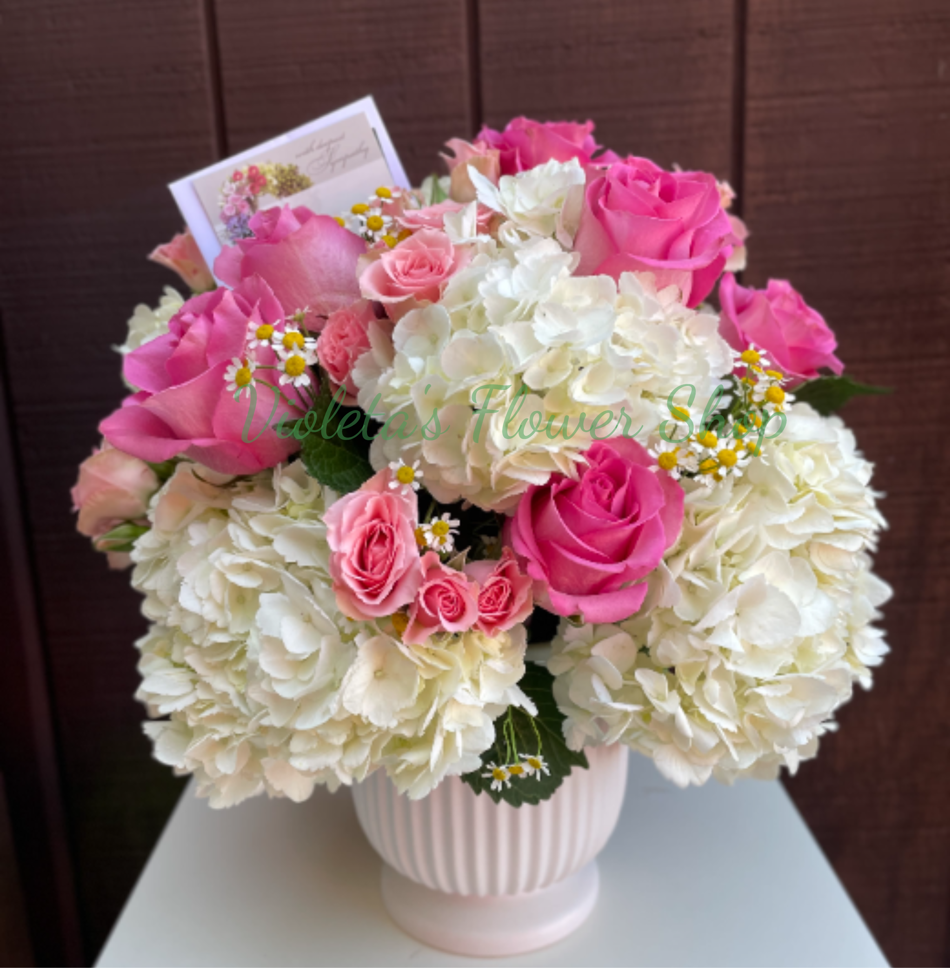 Roses and Hydrangeas Arrangement - Bright pink and blush roses rise above billowy white hydrangea clusters in this Roses and Hydrangeas arrangement, a cheerful get-well and thinking-of-you bouquet that reads as both romantic and comforting. The soft pops of yellow-centered white daisy accents add a light, airy texture so the person you're sending it to will feel genuinely seen and uplifted-perfect for welcoming a new baby, saying congratulations, or brightening a recovery room.  The arrangement is gathered in a pale ribbed ceramic vase with a polished, classic look and comes with a greeting card for your personal message. Large creamy white hydrangeas form a lush, rounded base while bright pink and blush roses give height and visual contrast; tiny white-and-yellow daisy-like flowers are tucked throughout for delicate detail. Sized to sit on a bedside table or mantel, it offers long visual impact and easy care-keep stems in fresh water and out of direct heat for best longevity.