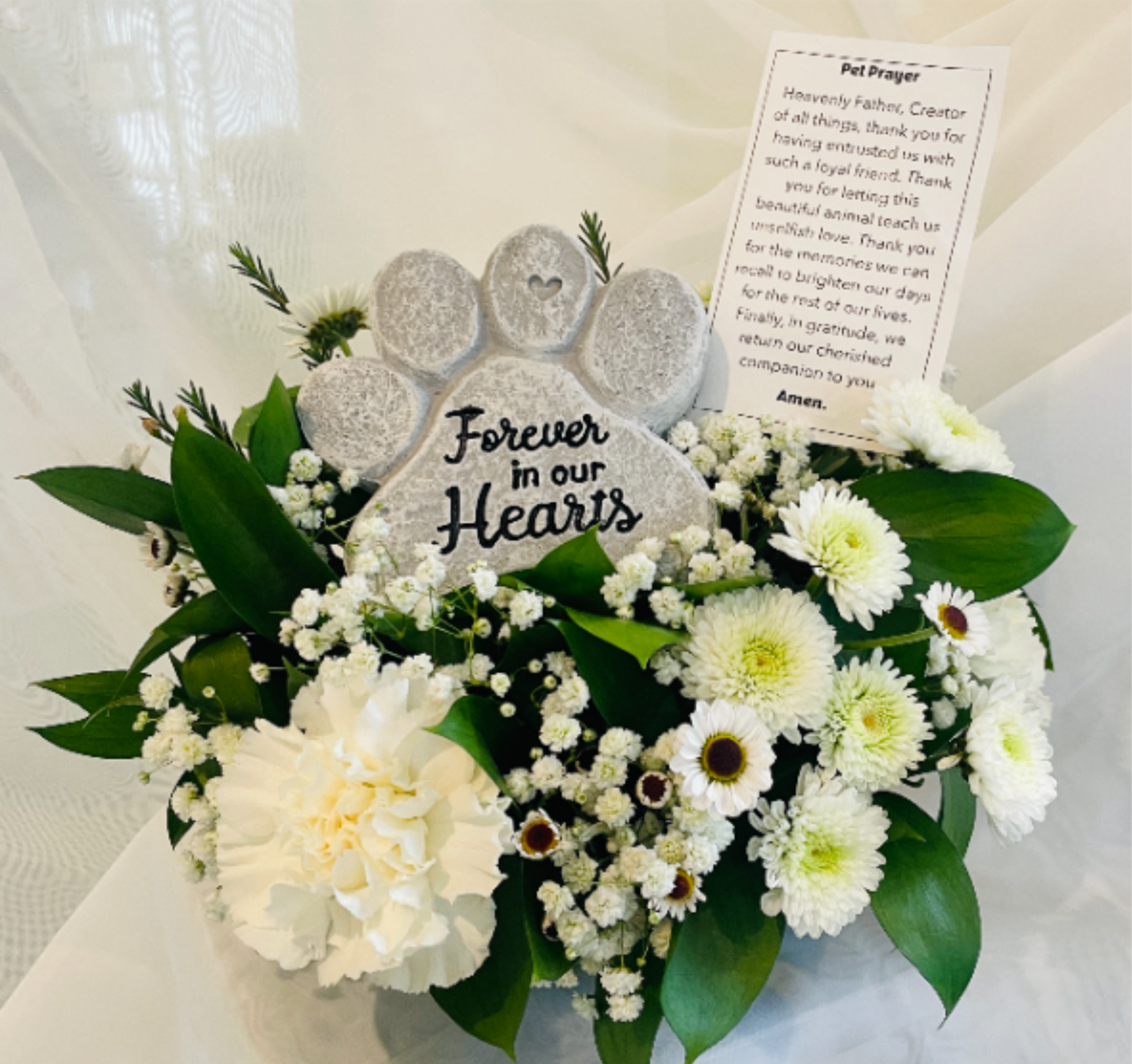 Gentle Paw Arrangement - A paw-print memorial stone reading "Forever in our Hearts" sits at the heart of this pet sympathy arrangement, creating an immediate focal point that feels personal and comforting. Designed as a pet memorial piece, the Gentle Paw Arrangement is the kind of tribute that helps the person you're thinking of feel seen and remembered during a time of loss. Perfect for honoring a beloved pet at a memorial table, garden or graveside, this arrangement reads as serene and respectful rather than celebratory. Size and delivery details are available at checkout; these flowers are arranged for immediate display and should be kept in a cool spot with fresh water as recommended on the included care card.