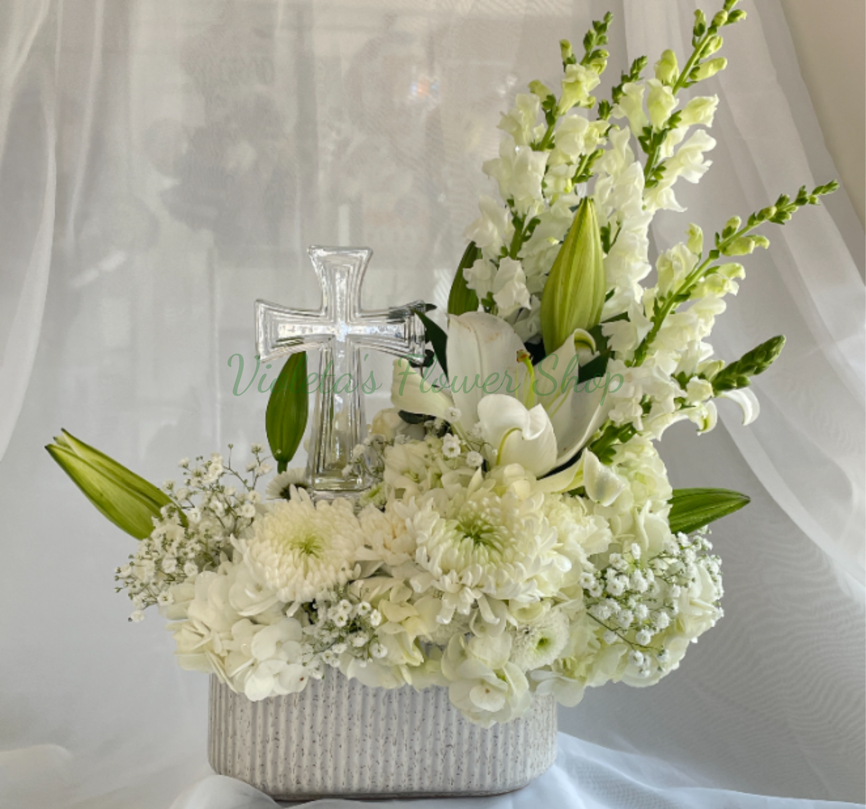 Heartfelt Sympathy Arrangement - A serene, upright sympathy arrangement centered on pure white lilies and a clear glass cross-designed to convey reverence and comfort for memorial services and church gatherings. This tall sympathy arrangement places white lilies, a white gerbera, daisies and pale snapdragon spikes in a clean white rectangular container (or white vase), creating a soft, monochromatic palette that reads as peaceful and respectful for funerals or moments of quiet remembrance.  