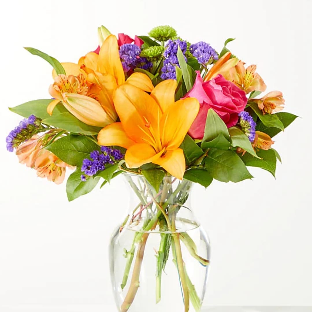Mixed bouquet of orange lilies, pink roses, and purple filler flowers in a clear glass vase