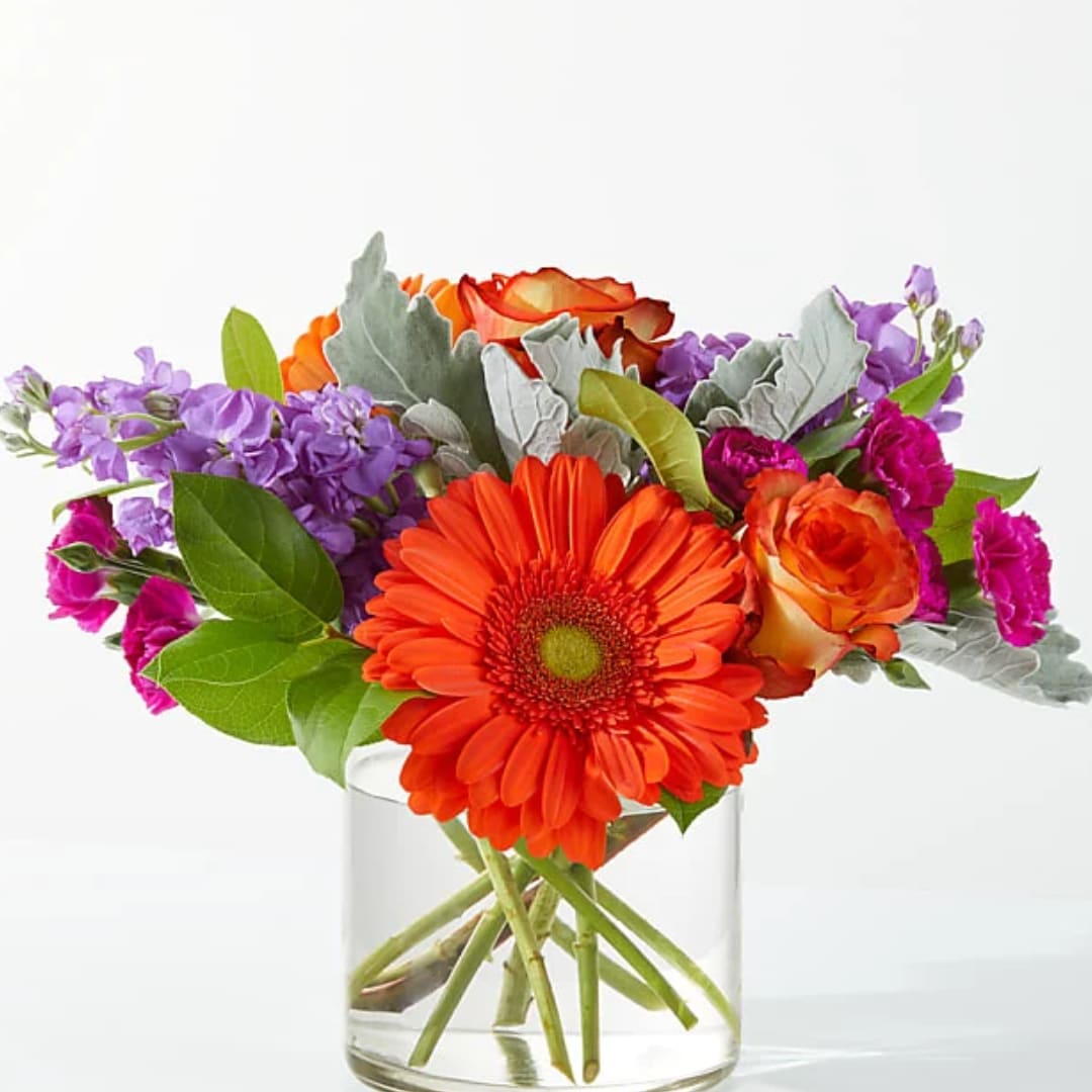 Orange gerbera daisies and roses in a clear glass vase with purple flowers