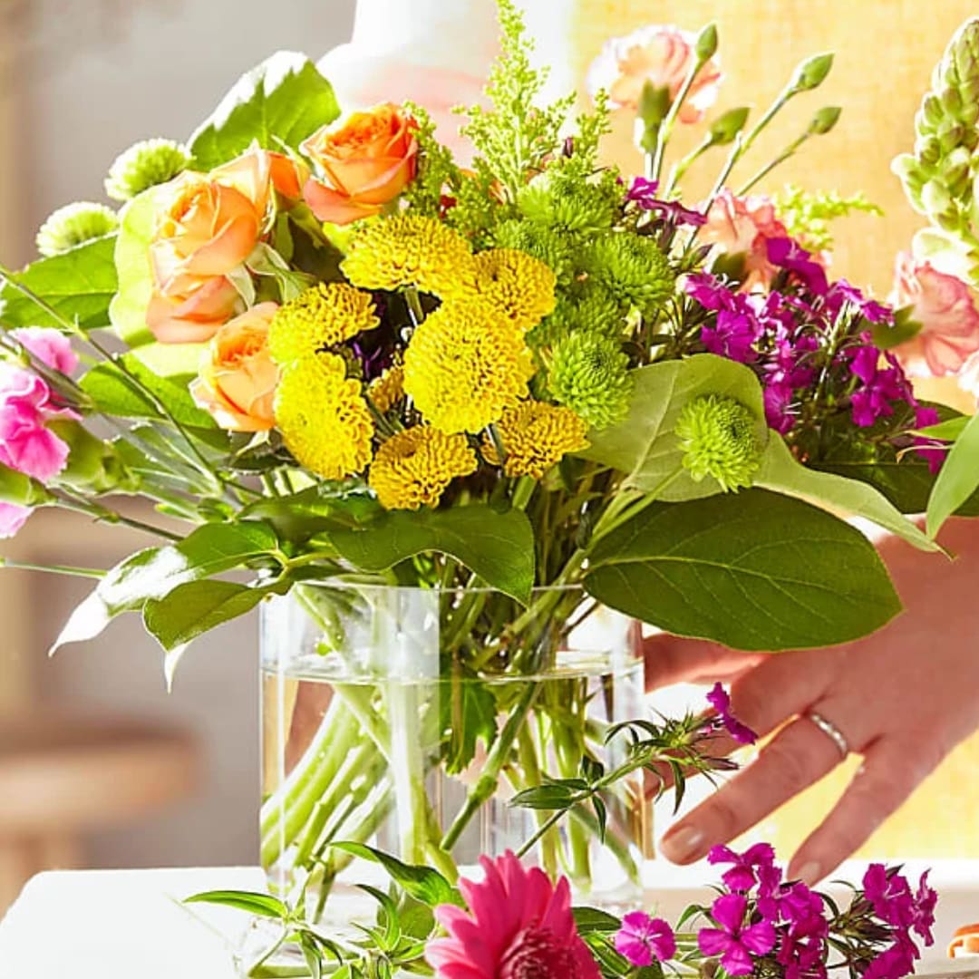 Mixed bouquet of orange roses, yellow mums, and purple flowers in a glass vase