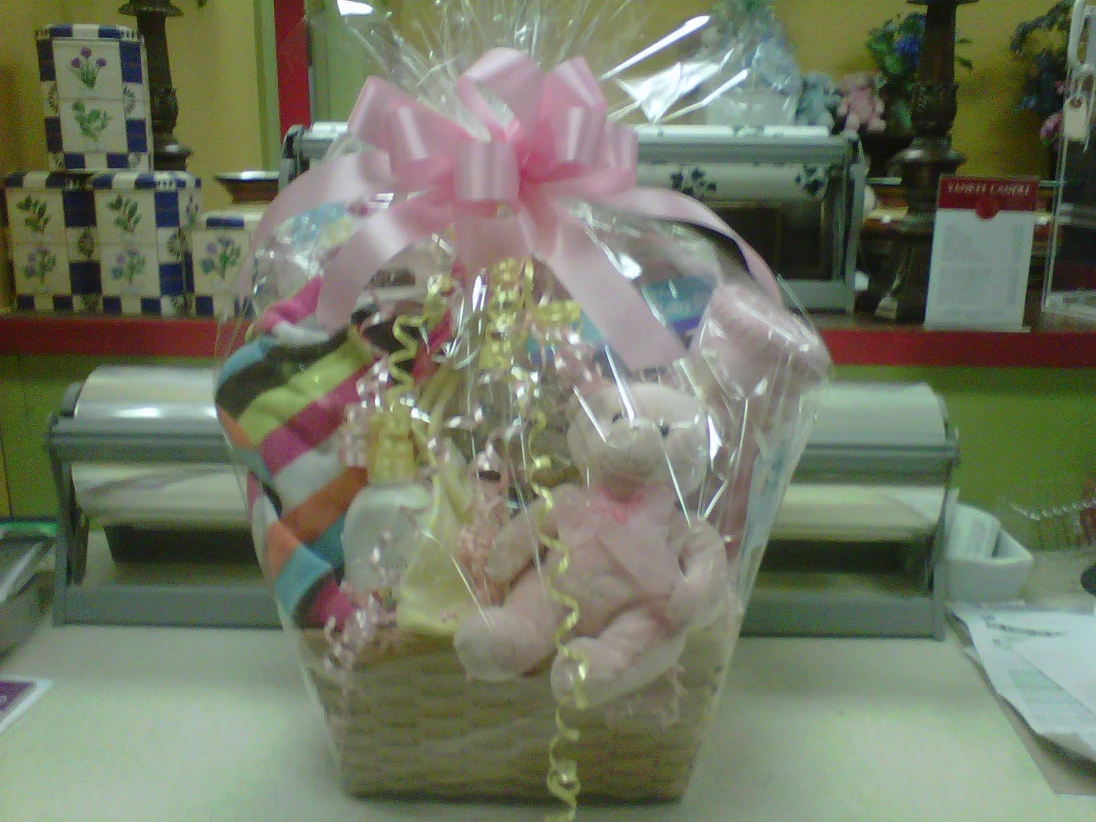 Gift basket wrapped in cellophane with pink bow, teddy bear, and baby items on a counter
