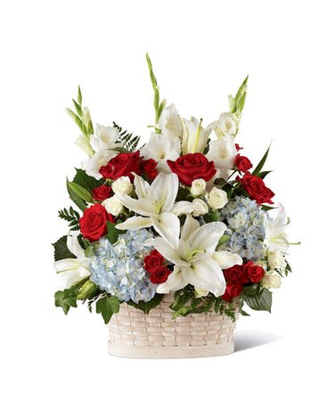 Patriotic basket arrangement with white lilies, red roses, and light blue hydrangeas in a white woven basket