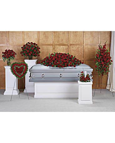 Funeral display with red rose casket spray, standing sprays, and heart wreath around a silver casket