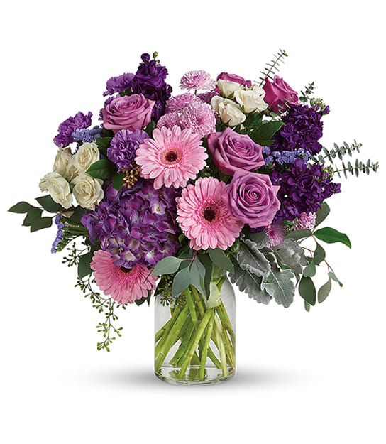 Mixed bouquet of pink gerbera daisies, lavender roses, and purple blooms in a clear glass vase