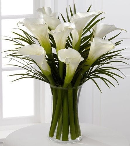 Tall arrangement of white calla lilies with long blades of foliage in a clear glass vase