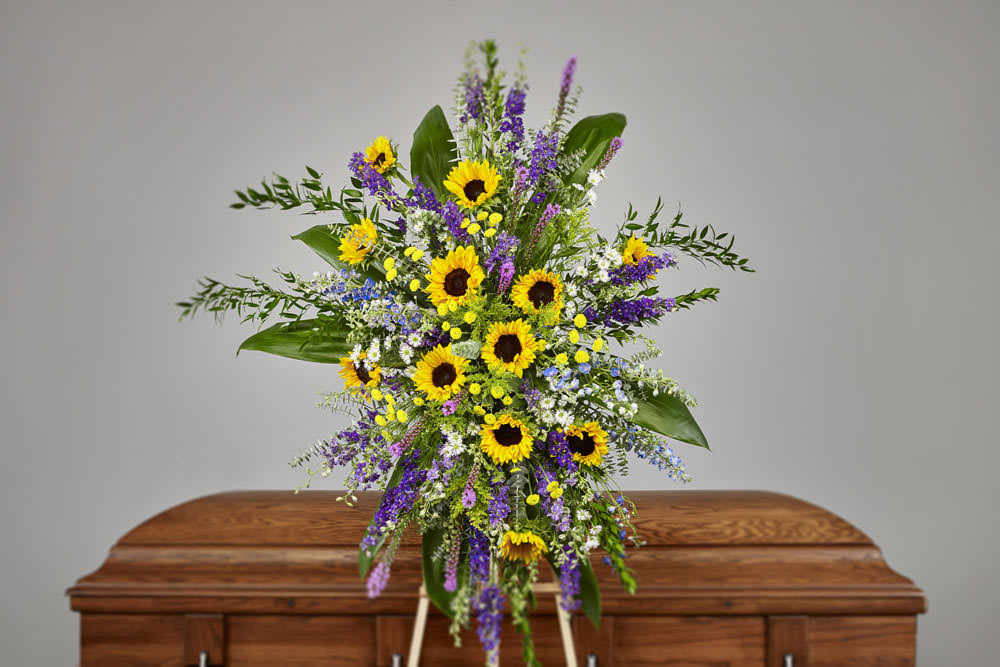 Standing spray of yellow sunflowers and purple flowers in front of a wooden casket