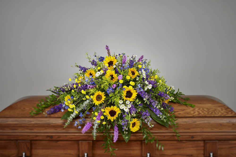 Bright casket spray of yellow sunflowers and mixed purple and white flowers on a wooden casket
