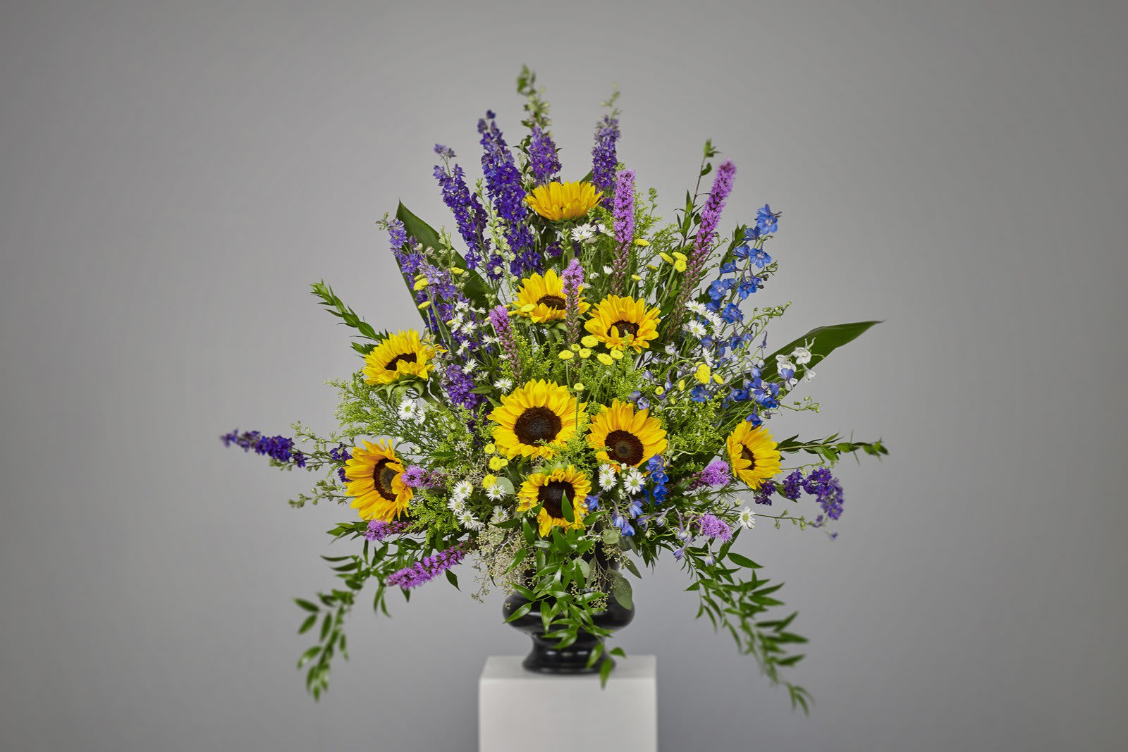 Tall arrangement of yellow sunflowers with purple and blue flowers in a black urn vase