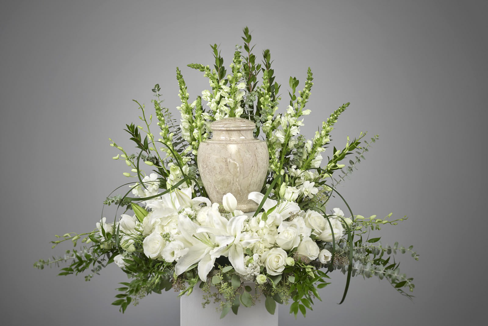 White lilies and roses arranged around a stone urn with tall white snapdragons behind.