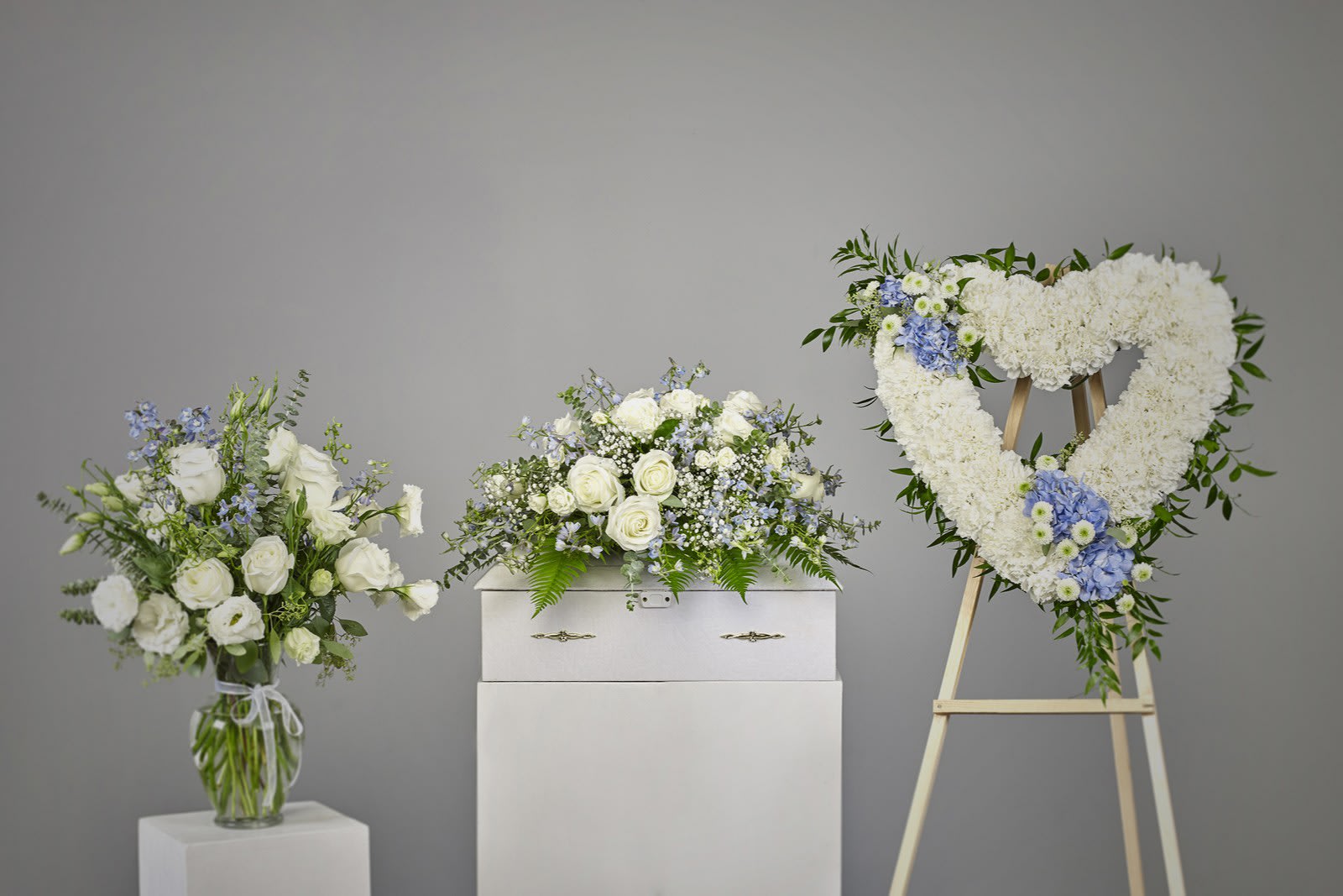 Sympathy set with white and blue arrangements and a heart-shaped wreath beside a white casket.