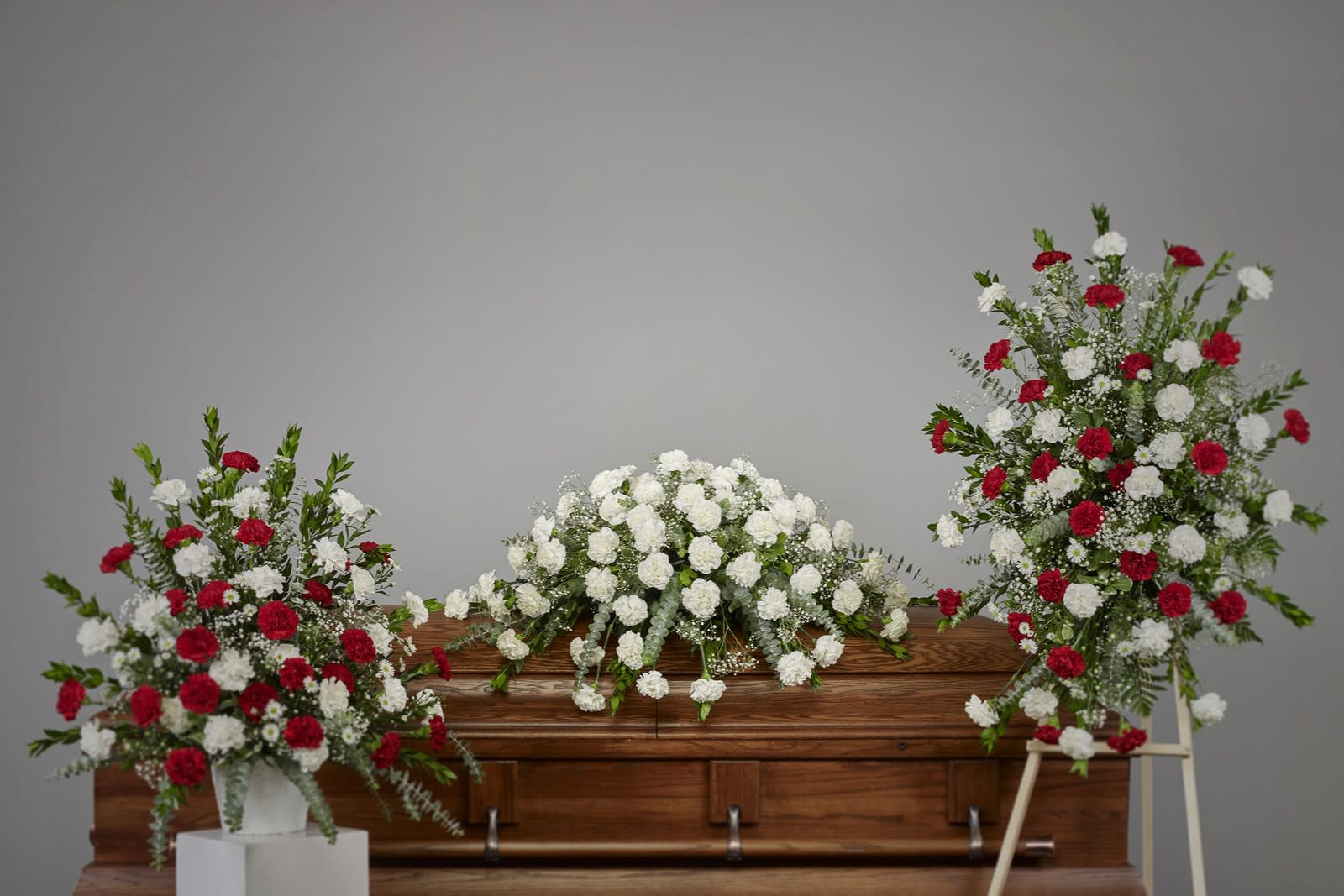 Red and white carnation sympathy set with casket spray and two standing arrangements beside a wooden casket