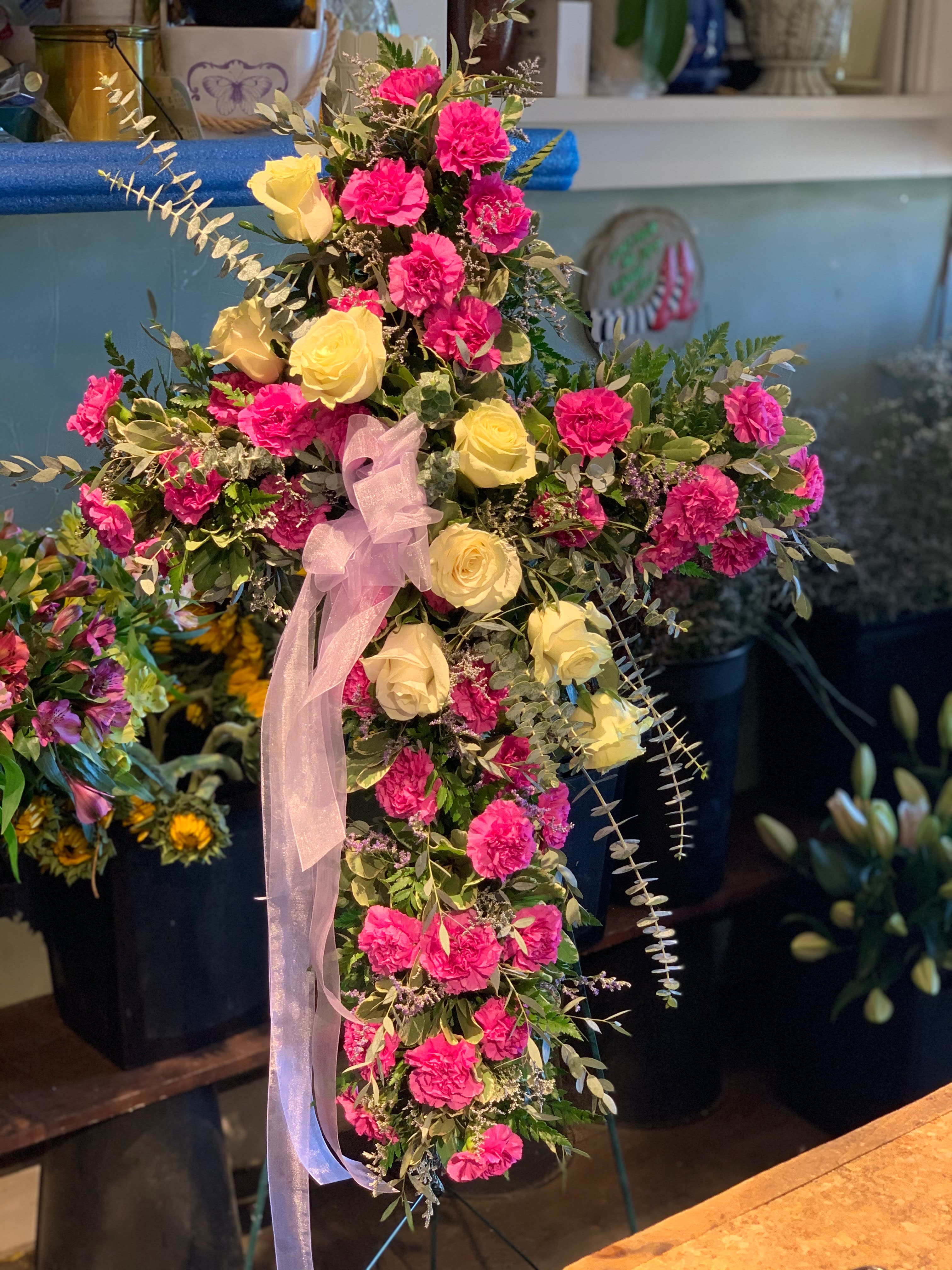 Standing cross spray of hot pink carnations and pale yellow roses with a sheer ribbon bow.