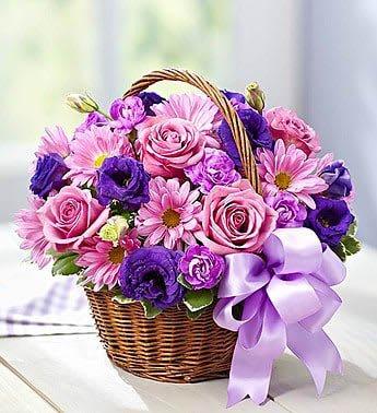 Pink and purple roses, daisies, and blooms arranged in a wicker basket with a lavender bow
